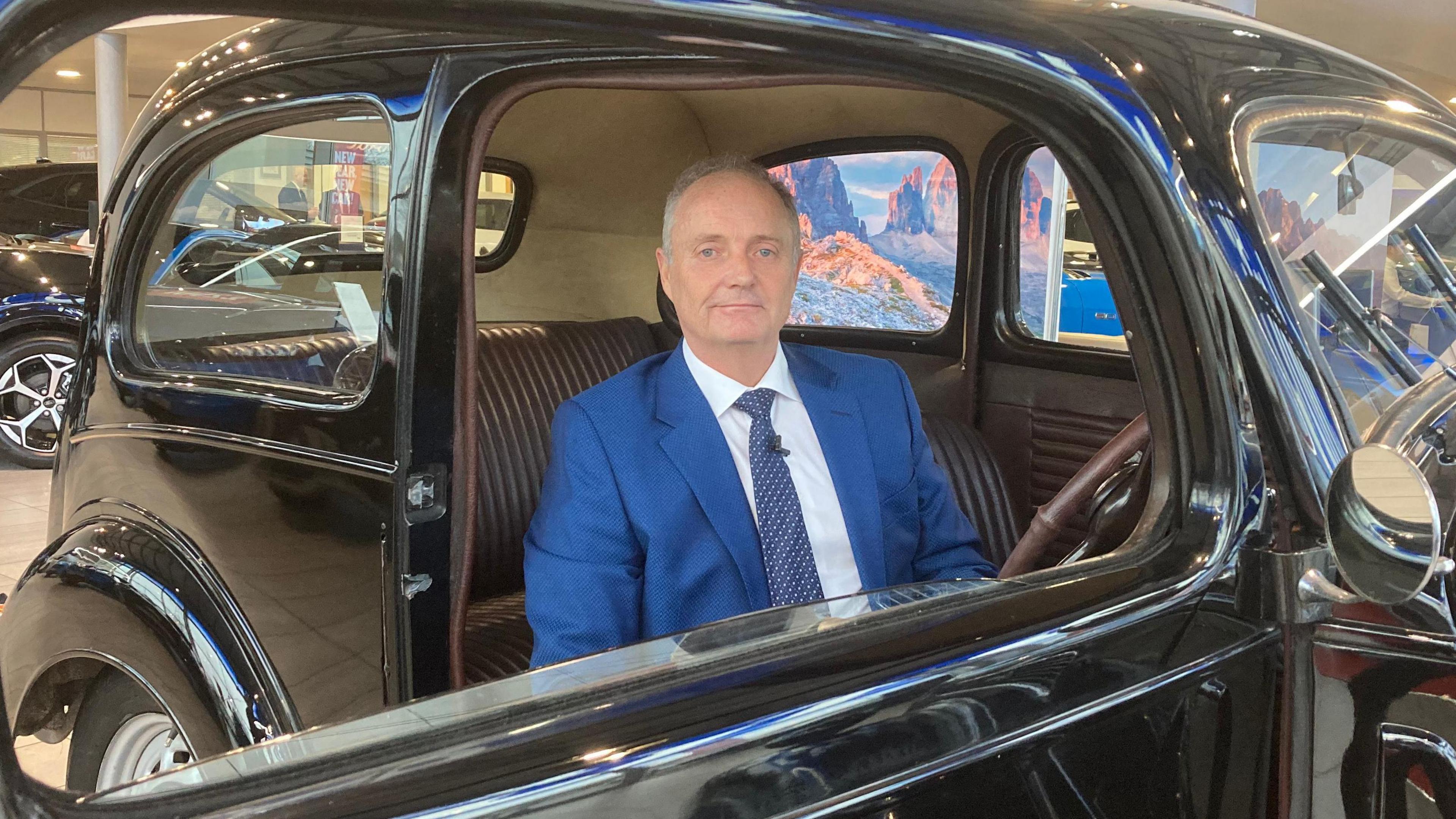 Paul Hendy the chief executive of Hendy Group sitting in the driver's seat of the 1949 Ford Anglia. He is wearing a blue suit and a blue tie with a white shirt.