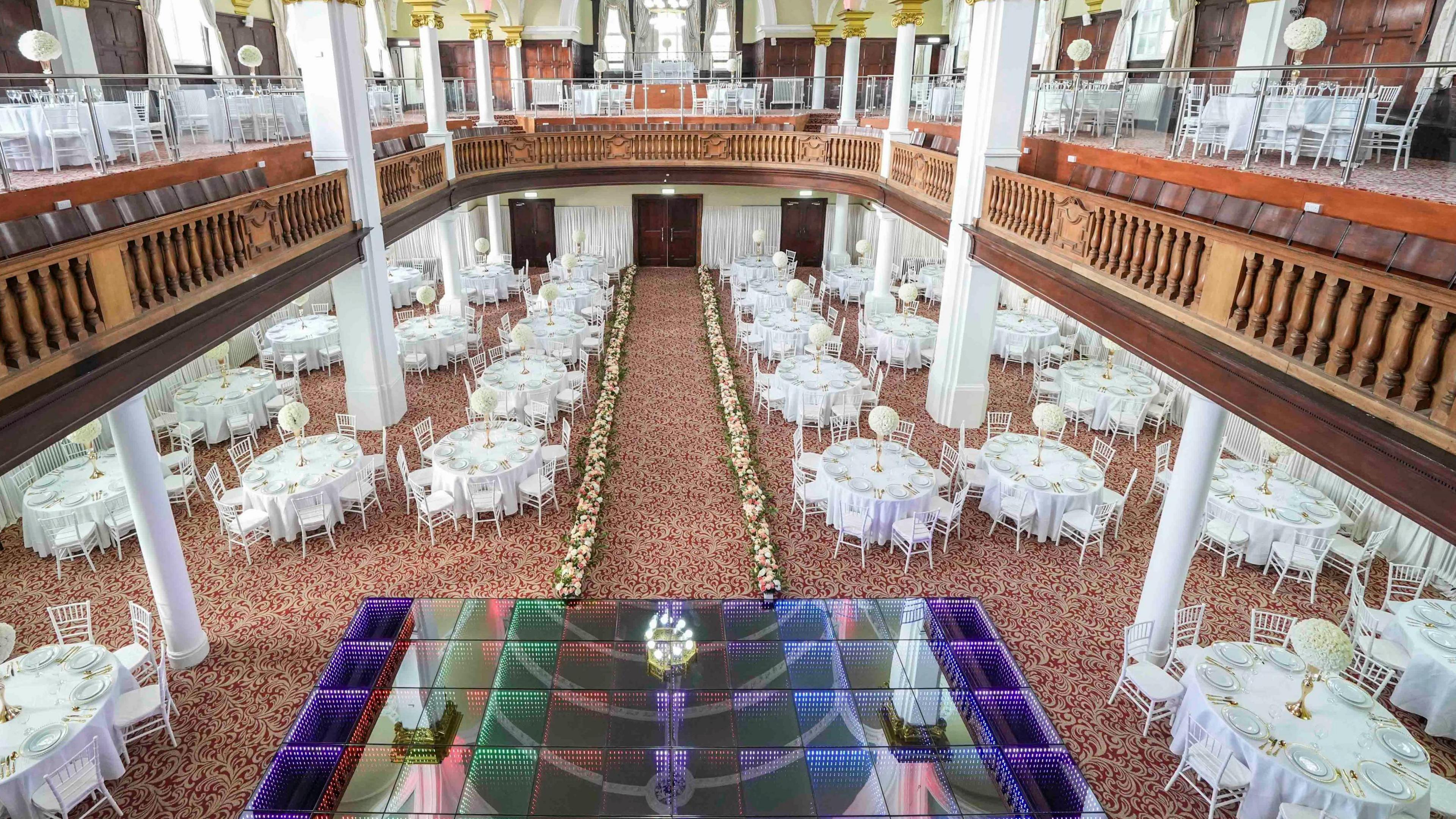 The inside of the building. White chairs are around the dance floor which is lit up. There are also chairs and tables on the upper floor which has a wooden and glass balcony. 