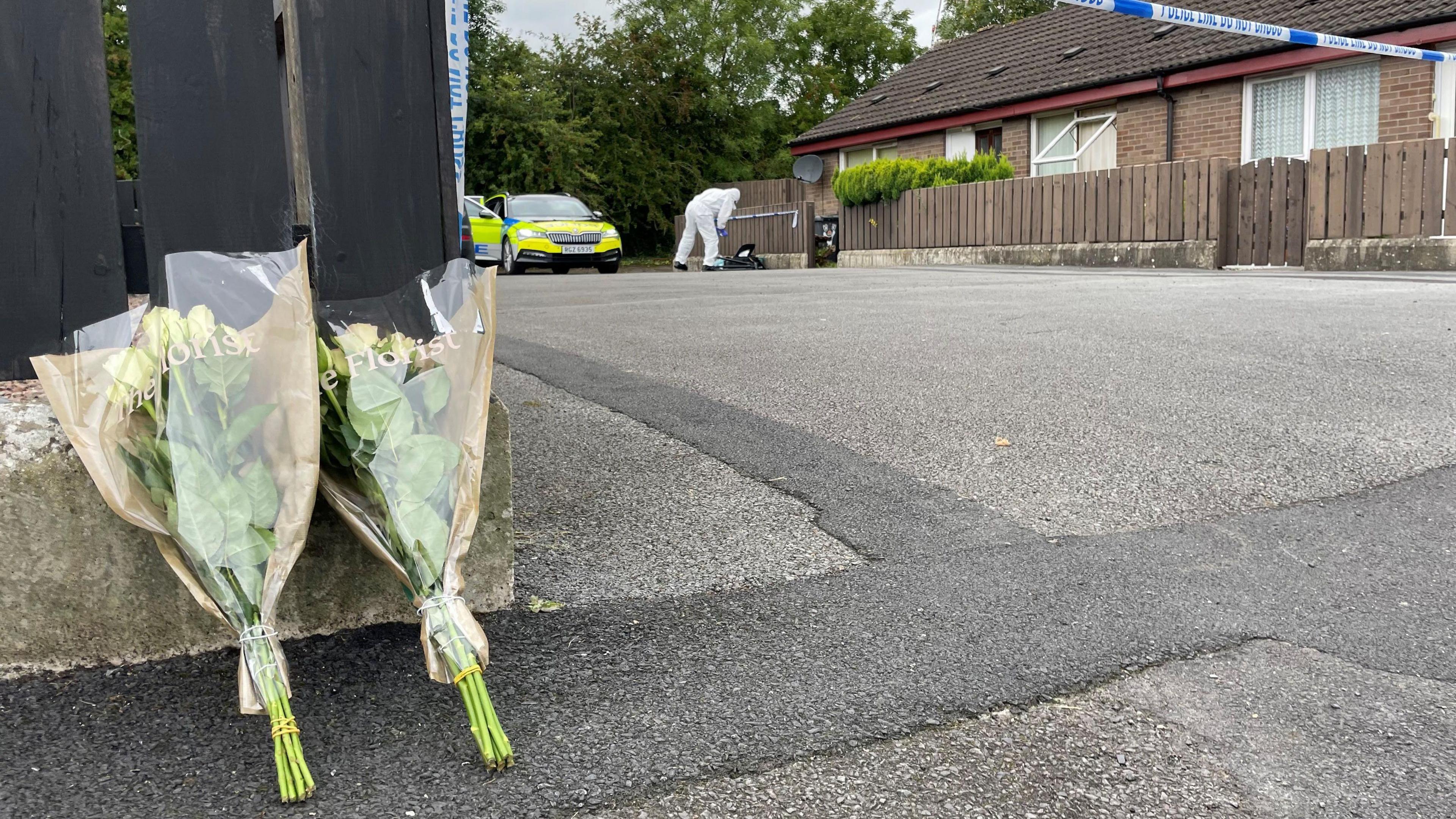 In the foreground two bouquets of flowers are propped up against a wooden fence. In the background there's a police car and a person in a forensic suit outside a red brick bungalow.