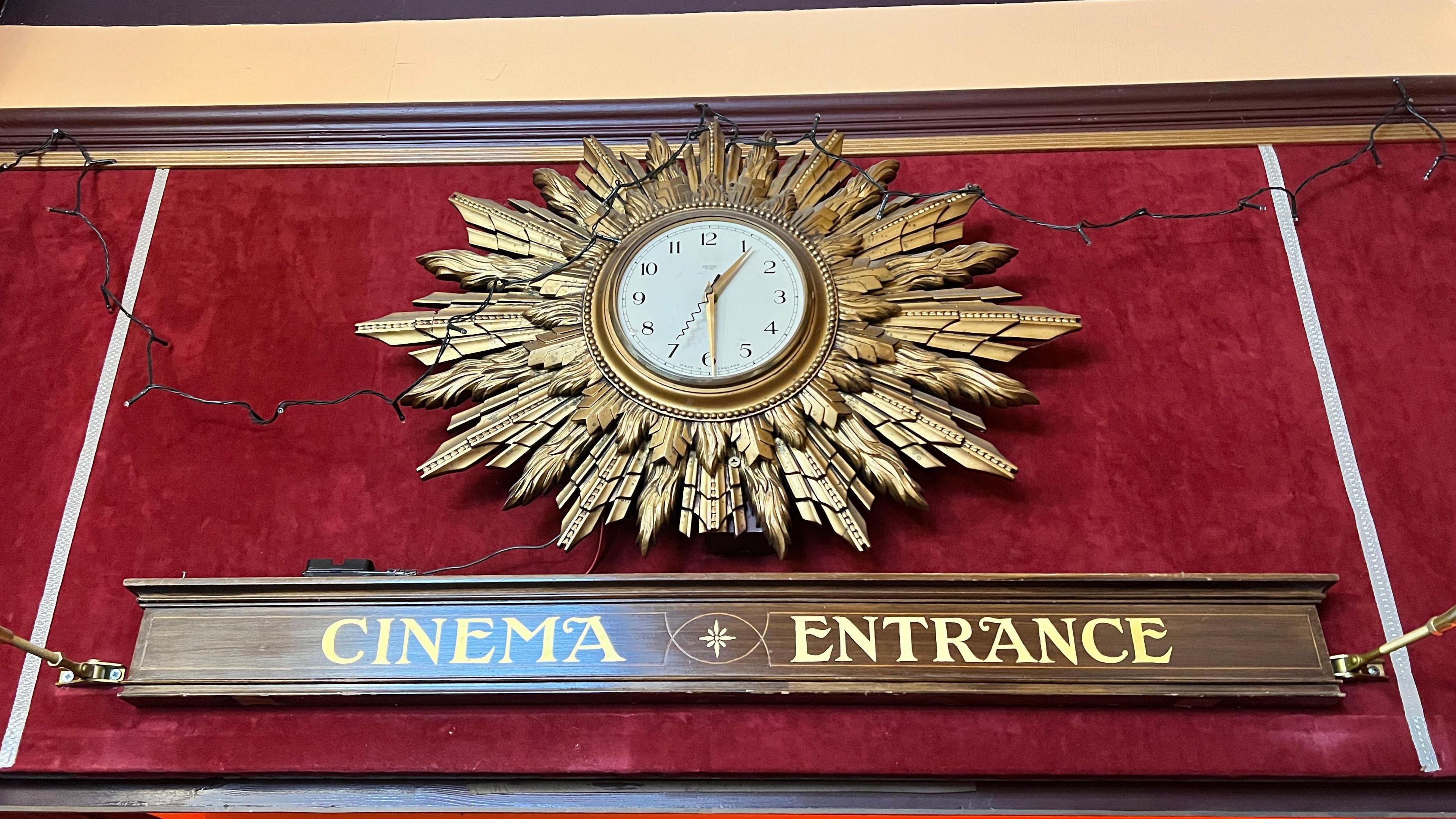 Above a wooden sign with gold lettering saying cinema entrance is a circular clock with gold coloured hands on a white background, with an elaborate golden surround looking like lots of feathers.