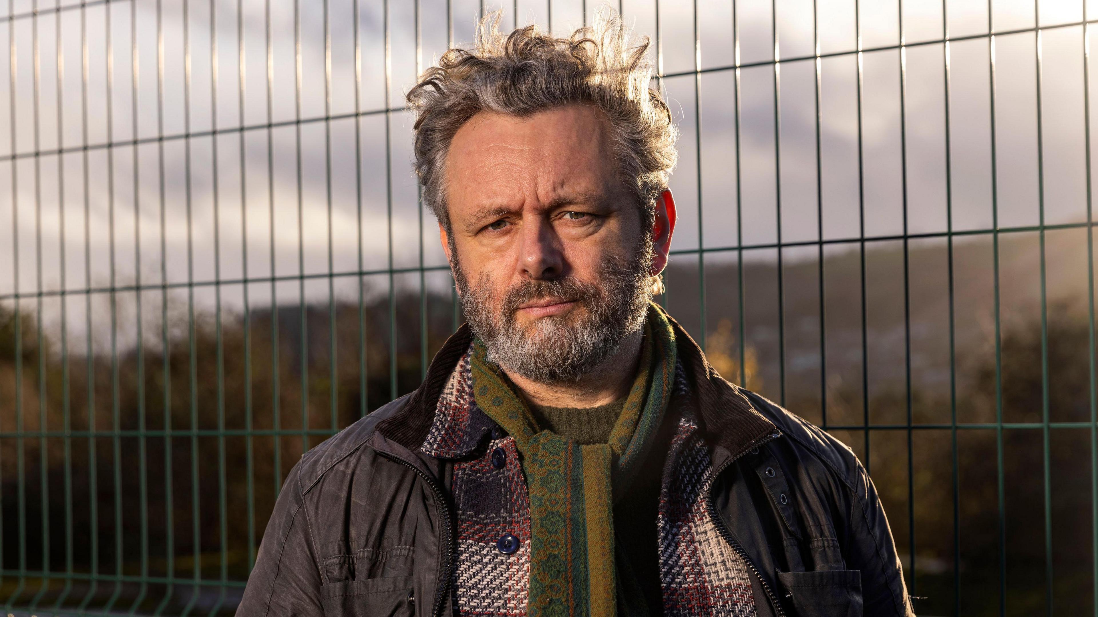 The image is a close outdoor portrait taken near a wire-mesh security fence. Michael Sheen stands facing the camera, dressed for cool weather in several layers, including a dark field jacket, a checked wool coat and a patterned green scarf. The fence fills much of the background, while distant hills and trees are softened by shallow depth of field. Golden sunlight from a low angle adds warmth to the scene and emphasises the textures of the scarf, coat and fence.