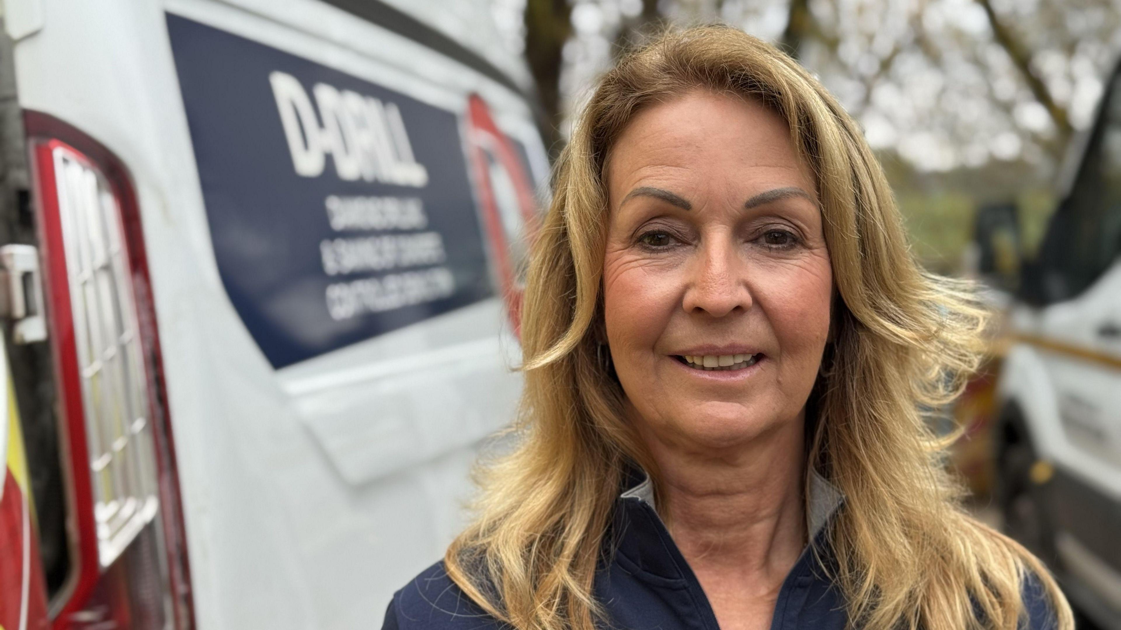 Julie White, a woman with blonde hair, is standing next to a white van. She is smiling while facing directly into the camera.