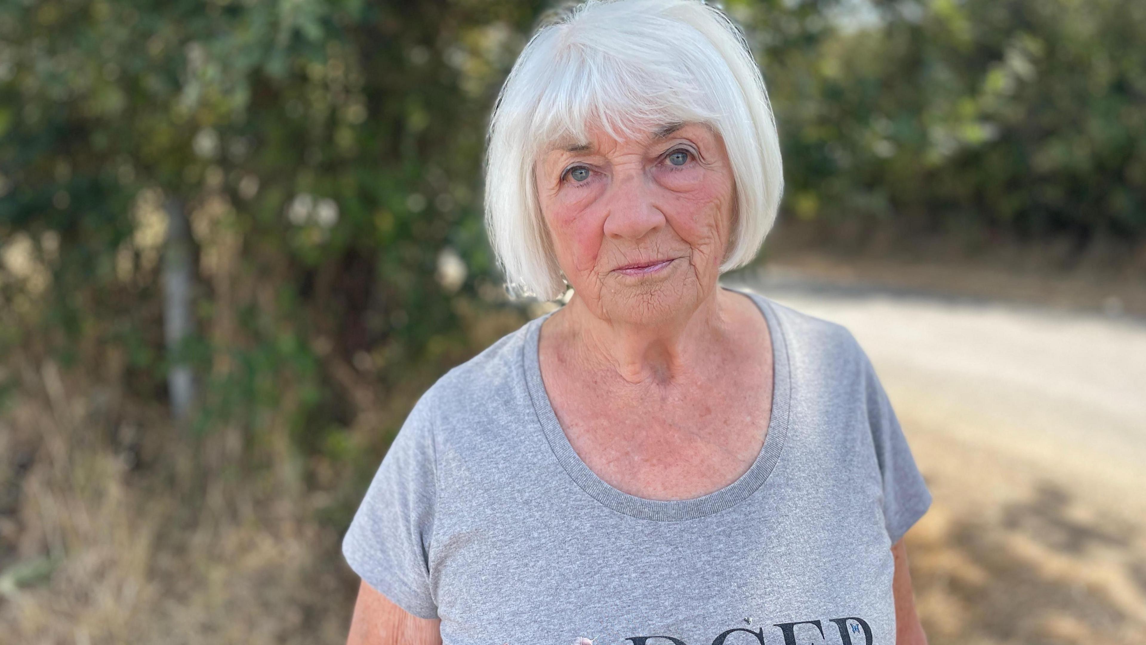 A woman with a silver bob haircut is standing outside wearing a grey T-shirt. She is looking directly at the camera with a serious expression on her face.