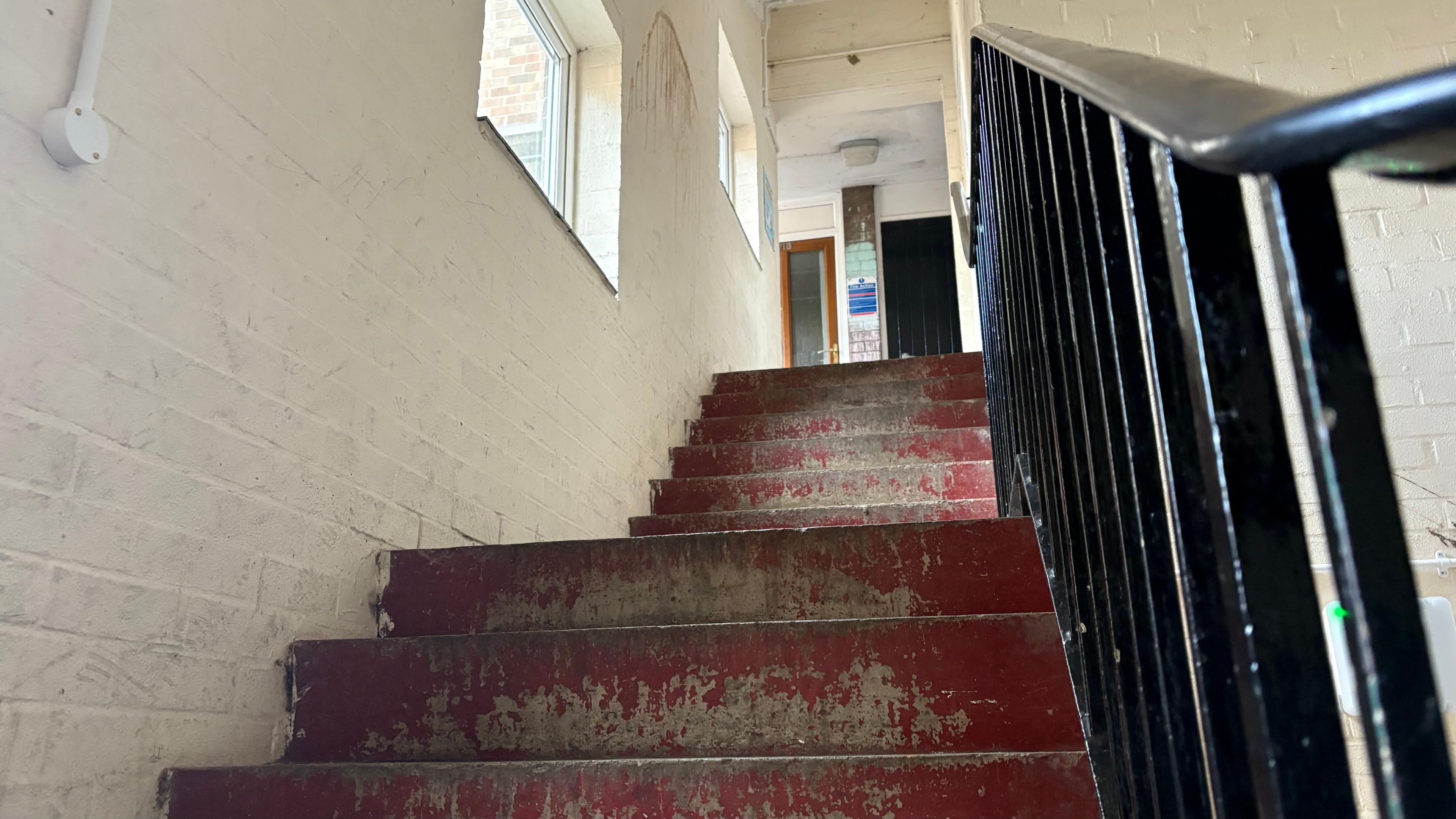 A view of the stairs in the block of council homes. They are dirty with the red paint coming off. The white wall next to the stairs is also dirty.