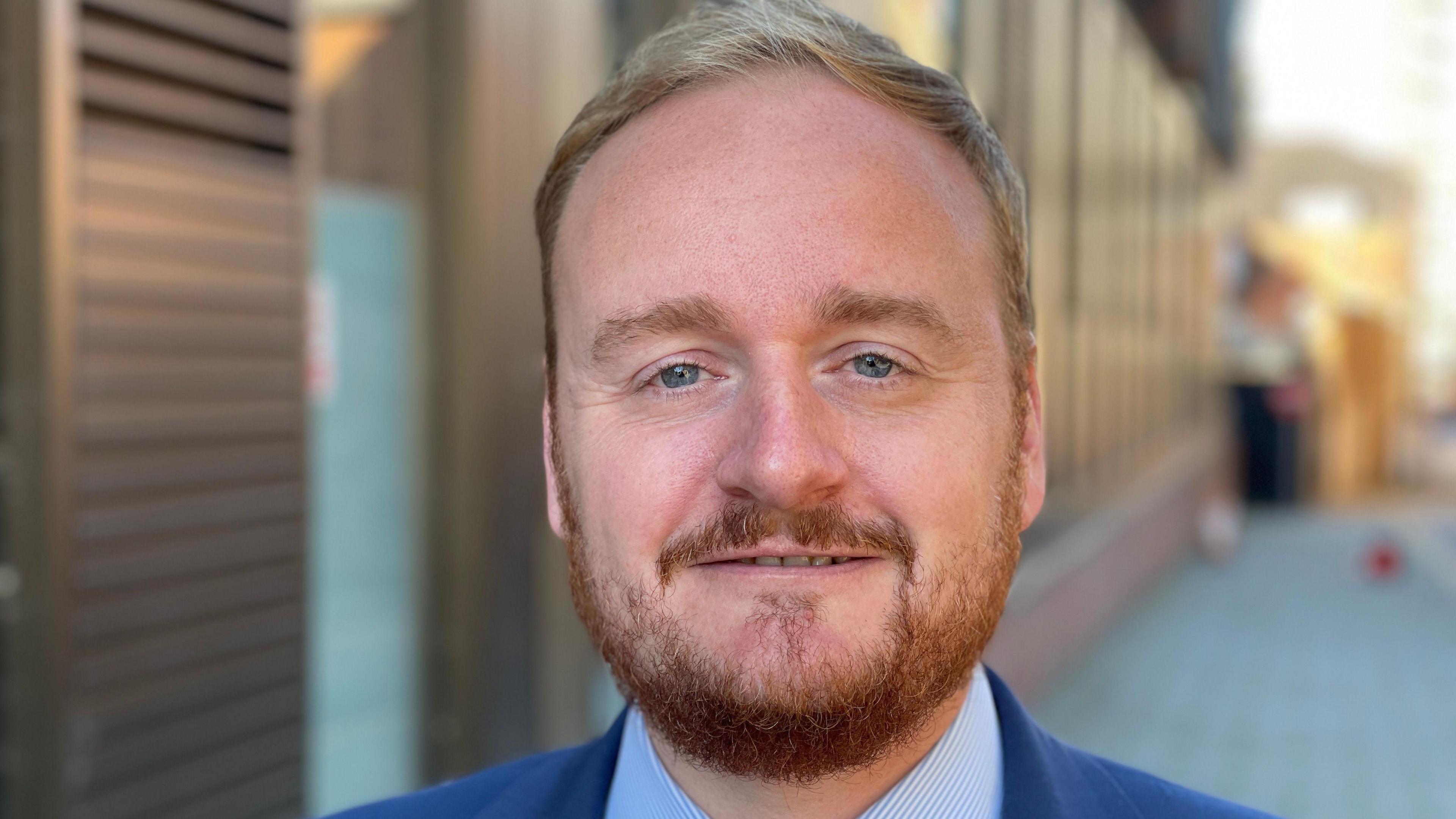 Adam Brown. A man with brown hair and a beard. He is wearing a blue suit