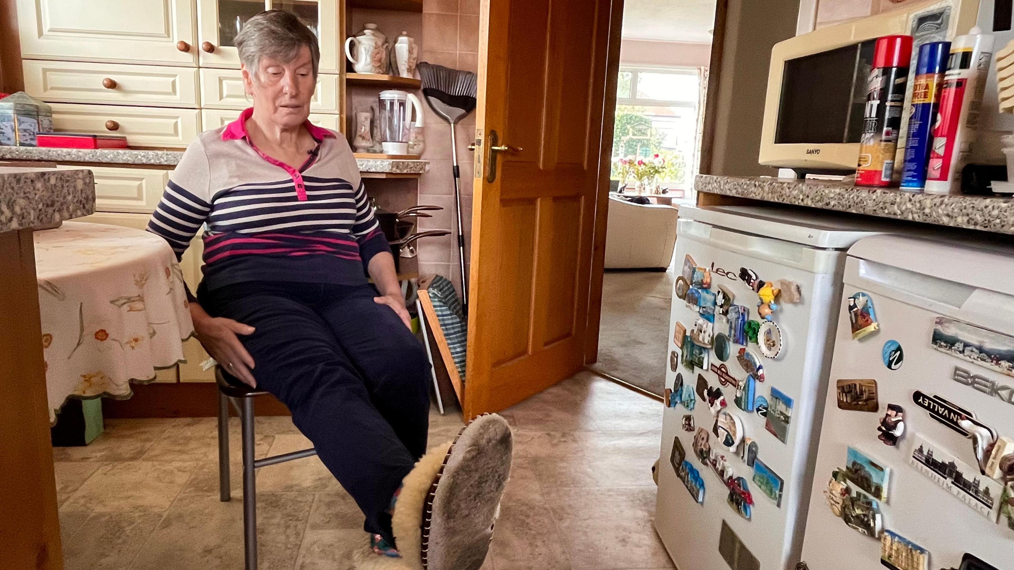 A woman sitting on a chair in her kitchen lifting her leg up. She is bending her recently operated-on hip