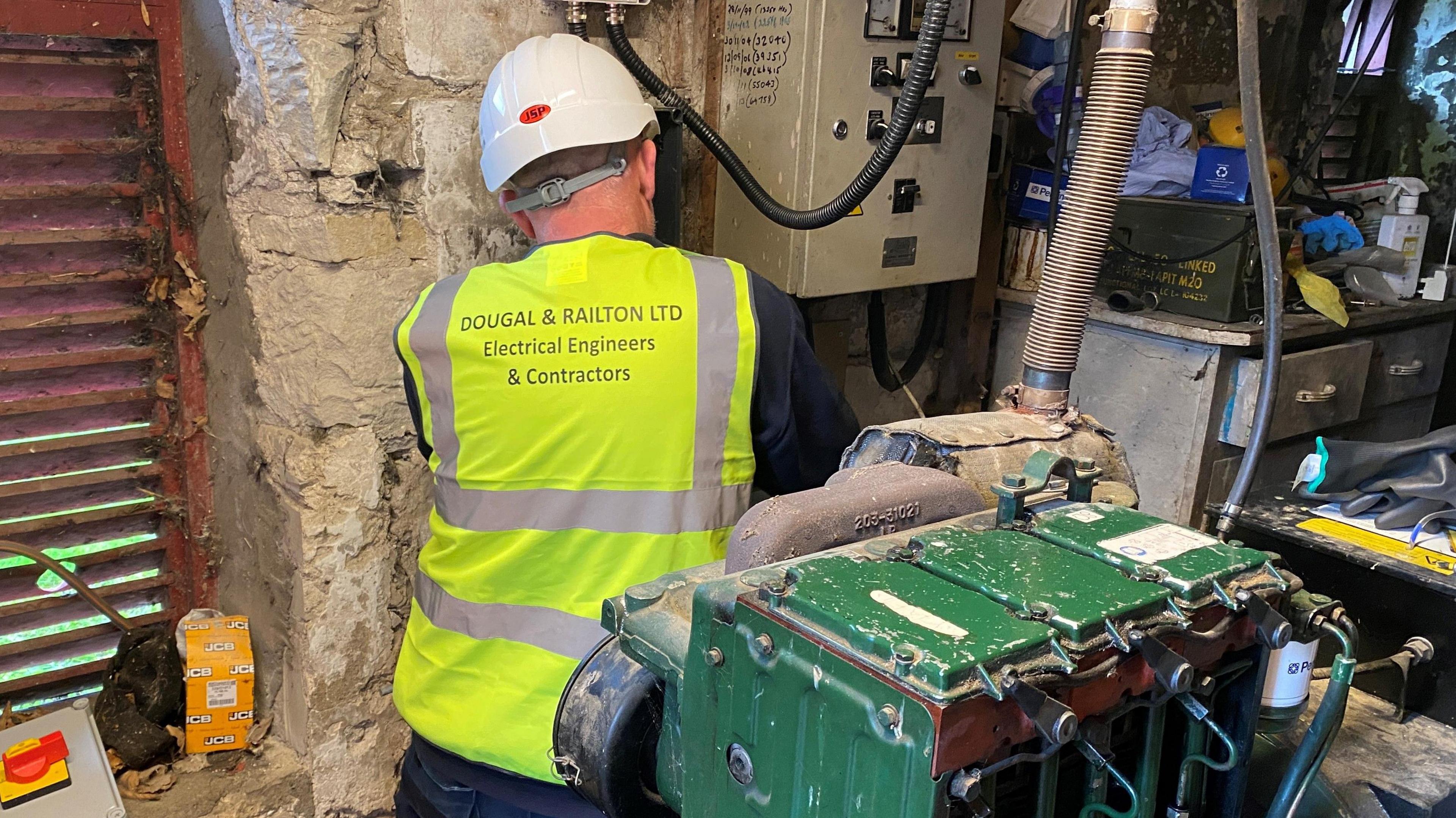 An engineer wearing a hi-vis jacket is working next to an electricity meter with a green generator behind him