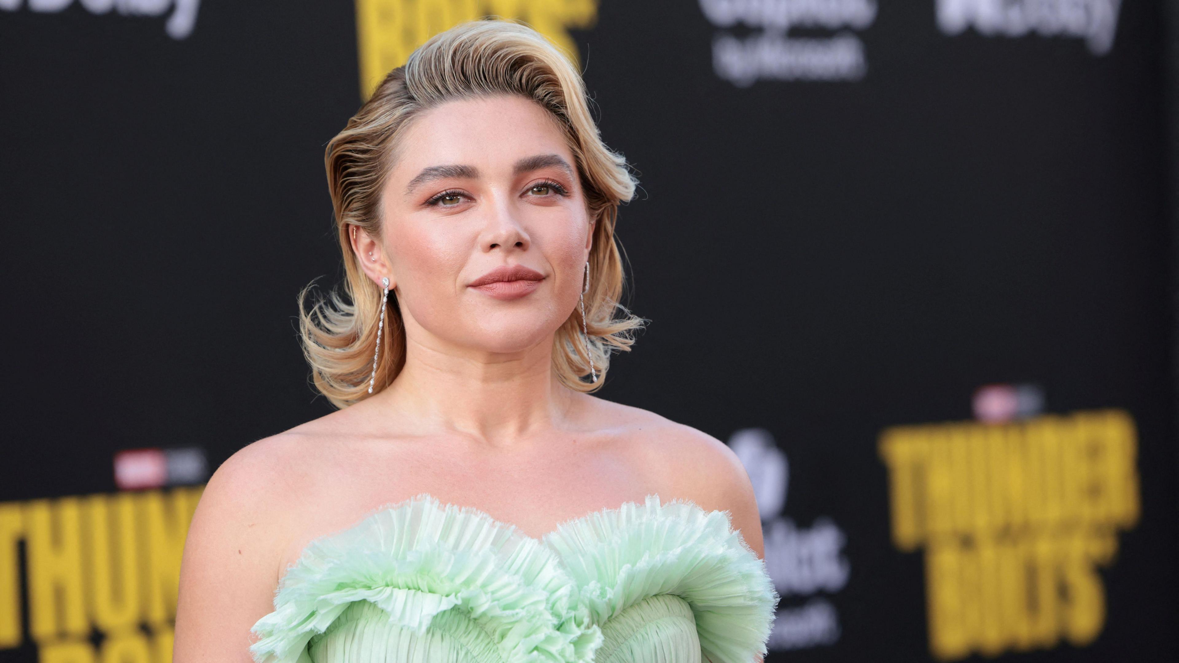 Florence Pugh, wearing a light green strapless dress and with her short blonde hair styled into a quiff, smiling at the camera against a black background.