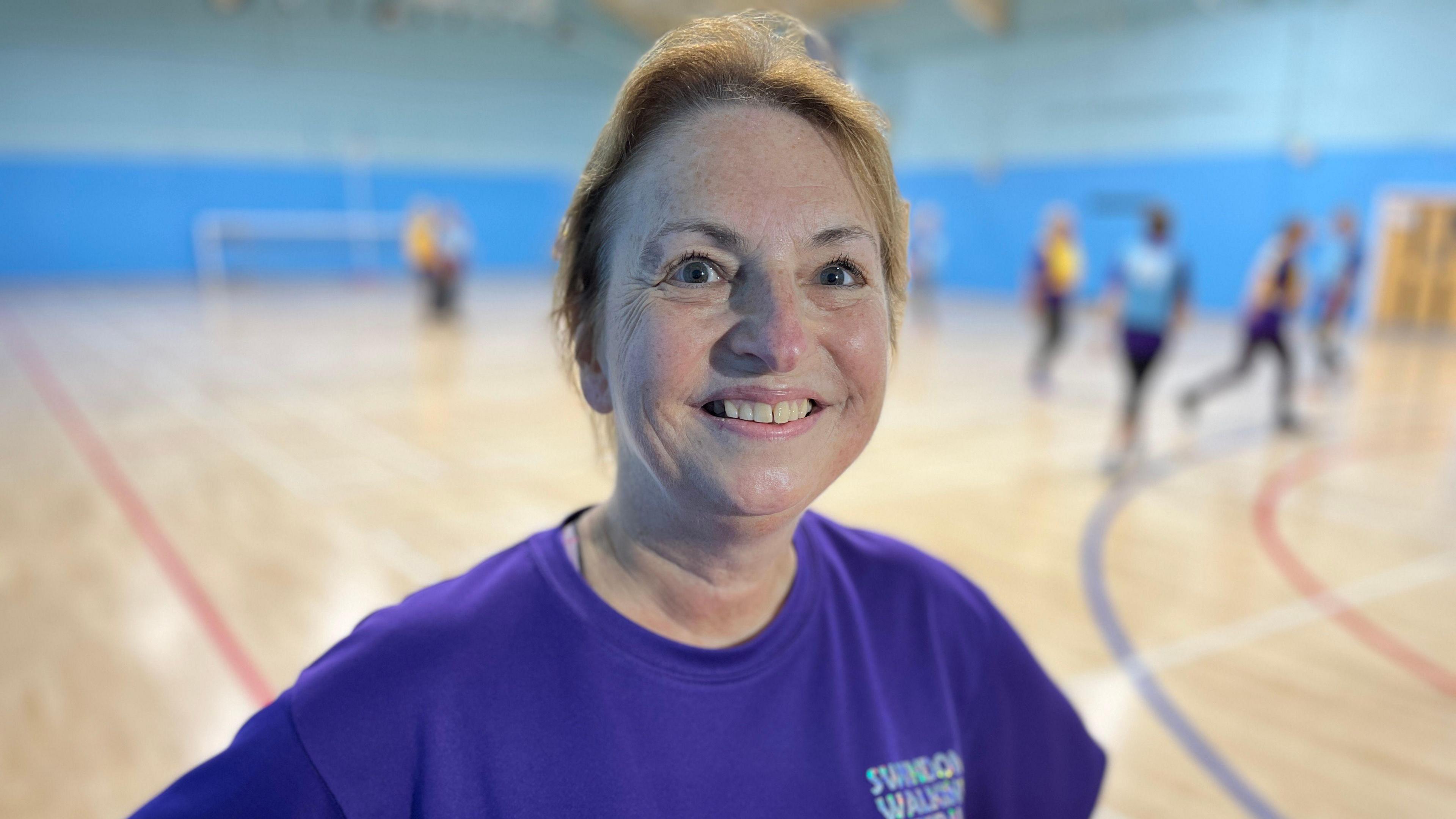 Nicki Sellars smiles into the camera. She's wearing a Swindon Walking Netball purple t-shirt. In the background women are playing walking netball.