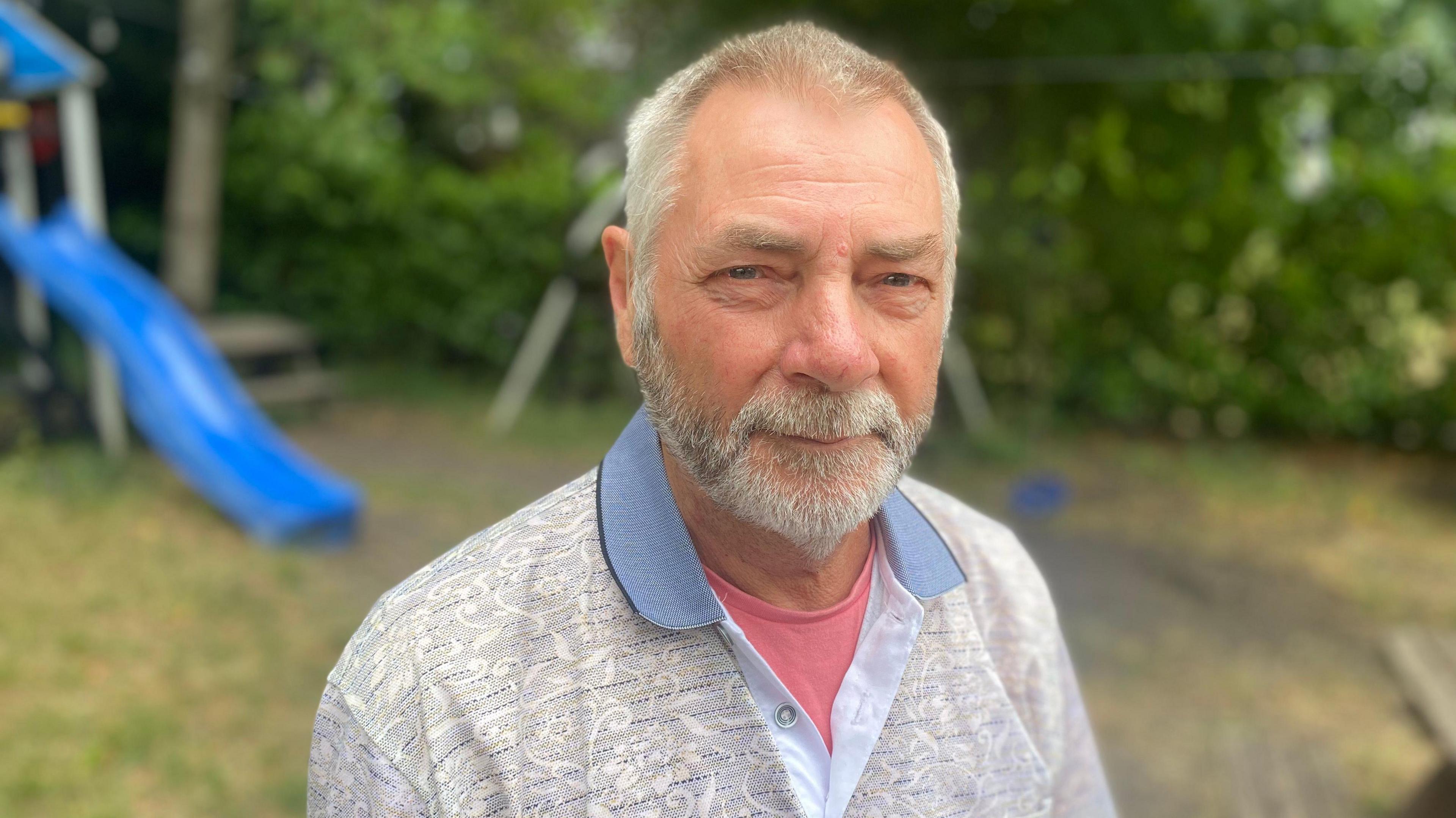 Mr Harris is smiling at the camera while outside. He has a grey beard and grey short hair. He is wearing a grey and blue top over a salmon pink T-shirt.