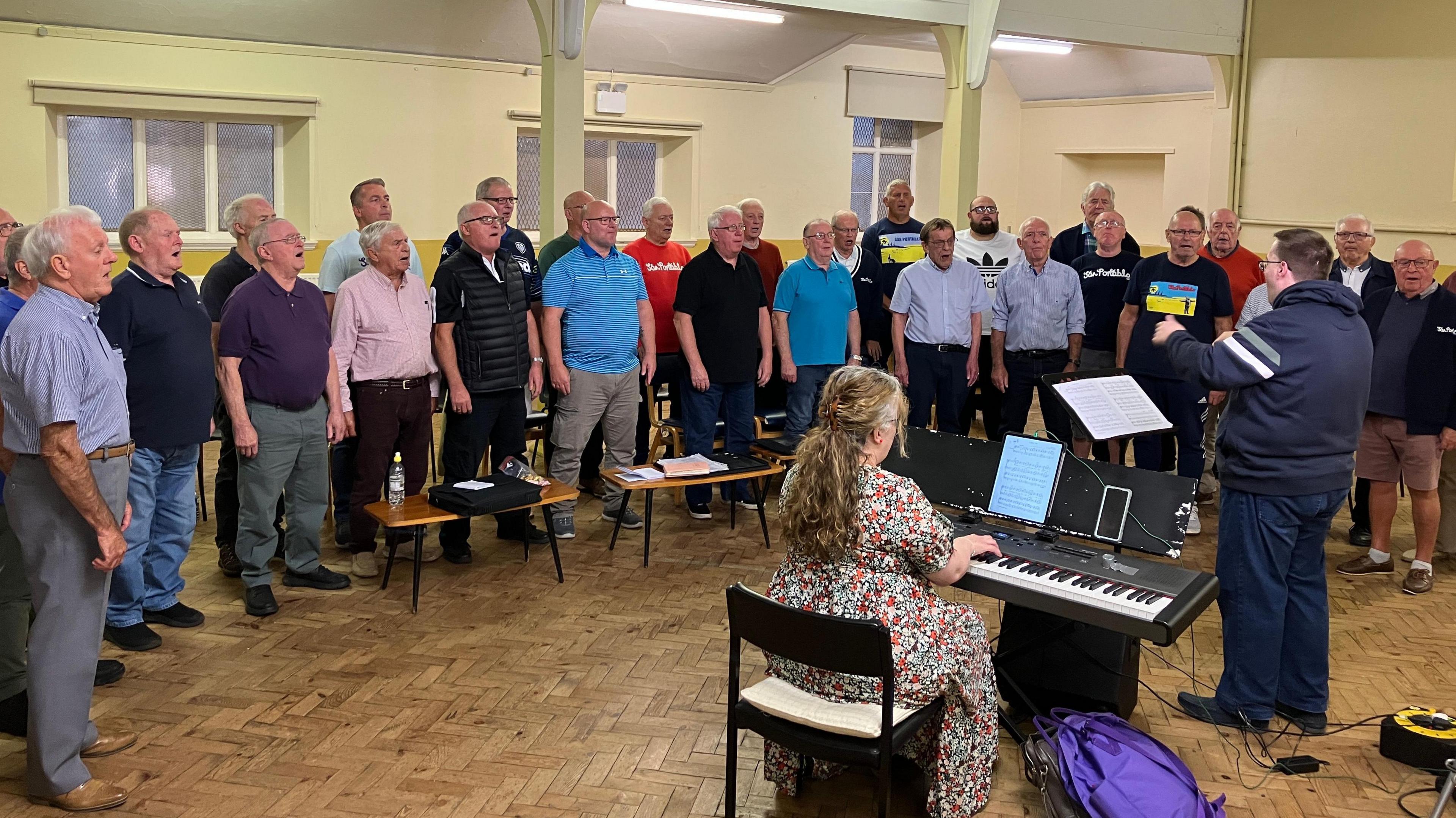 Male voice choir standing in a semi circle singing in a room which has cream walls and parquet flooring. In front of them with his back to the camera is the conductor and to his left is a female pianist who is wearing a floral dress.