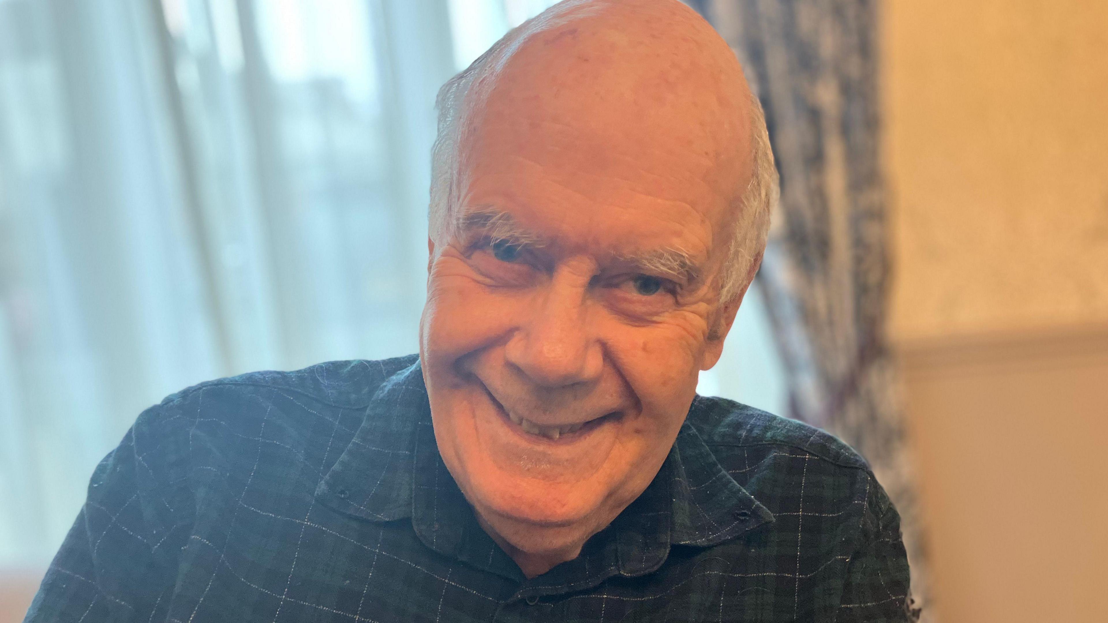 A man with white hair and wearing a green checked shirt smiling at the camera with a window behind him.