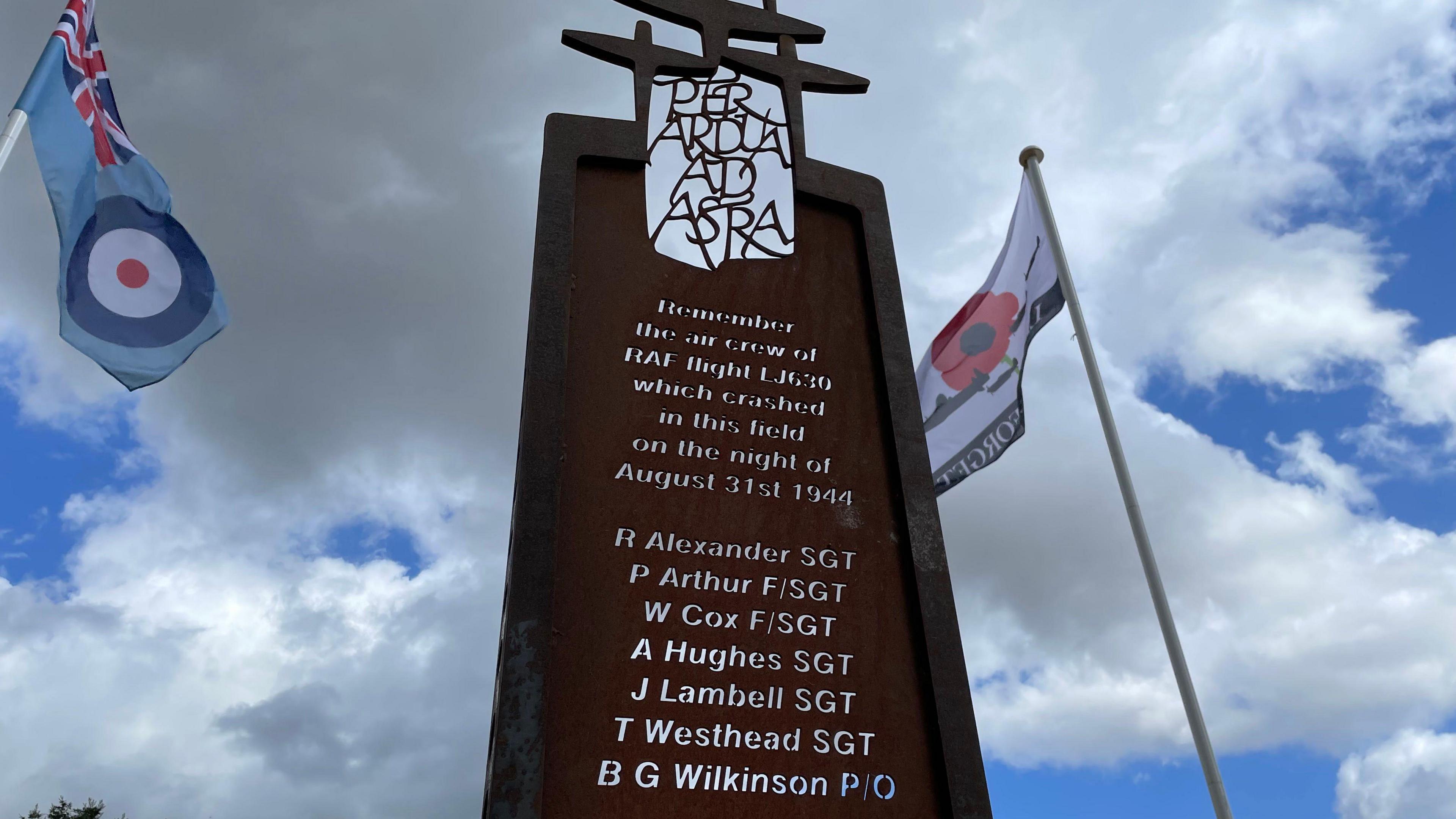 The memorial, a large upright piece of metal, roughly the size and shape of a door, with the dedication and names of aircrew cut through it to show the sky behind. It reads: "Remember the air crew of RAF flight LJ630 which crashed in this field on the night of August 31st 1944", with a list of the seven names underneath (as featured later in this story). Either side of the memorial, RAF and Poppy flags are flying.
