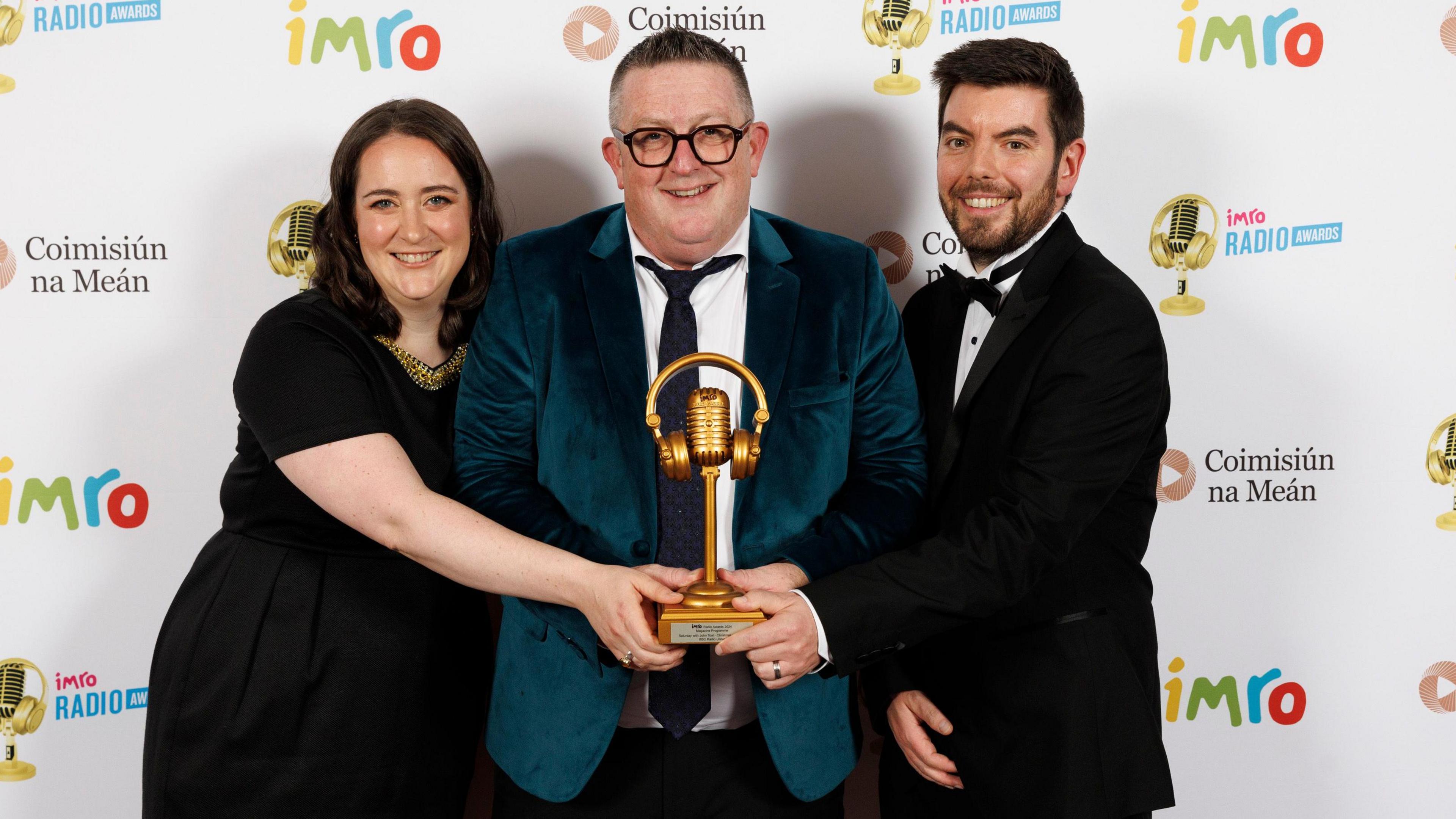 John Toal in the centre wearing a green/blue velvet jacket smiles as he holds a gold microphone IMRO award. On one side a woman with brown hair in a black dress reaches over to touch the microphone, on the other side a man in a tuxedo does the same