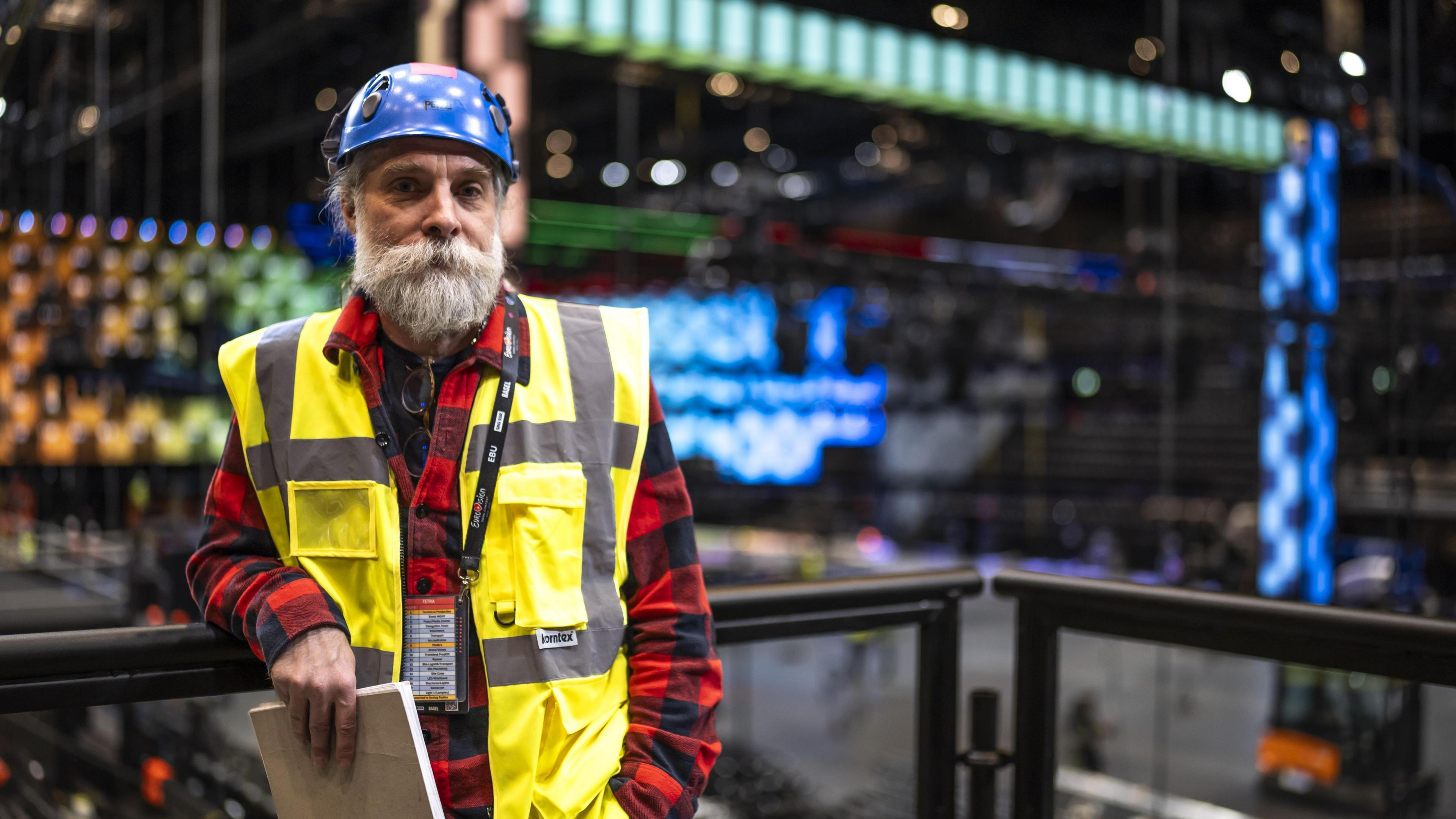 A man with a large beard wearing a flannel shirt, a helmet and a high-vis jacket poses for the camera as lighting infrastructure is erected behind him in a large stadium.