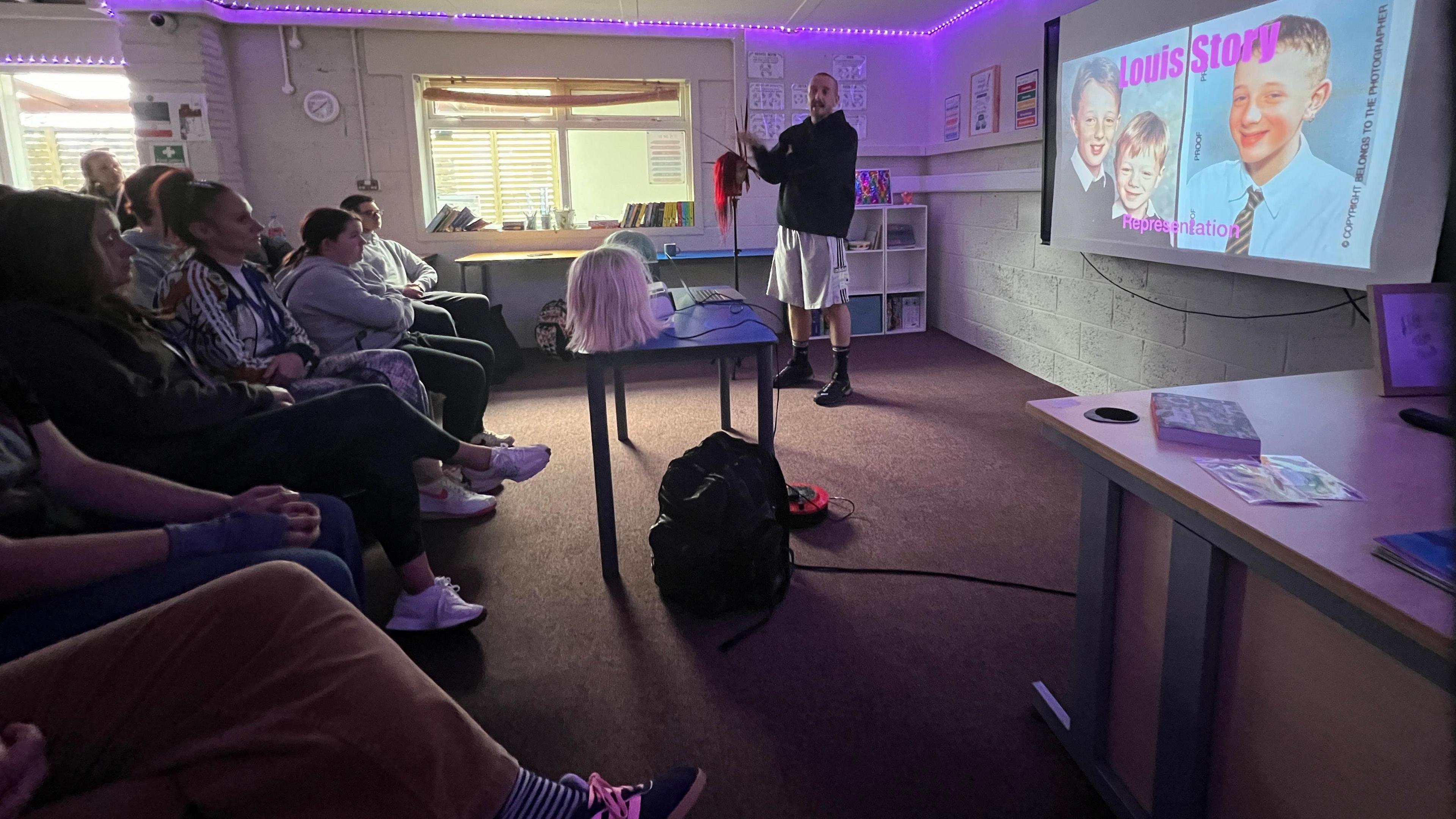 Louis stands in a classroom with the lights off and some purple fairy lights on around the ceiling. He is wearing a black hoodie and long white shorts and long dark socks. He's in the corner of the classroom giving a speech in front of staff and students. He's story next to a projector screen which has images of him as a school child on and reads Louis Story.