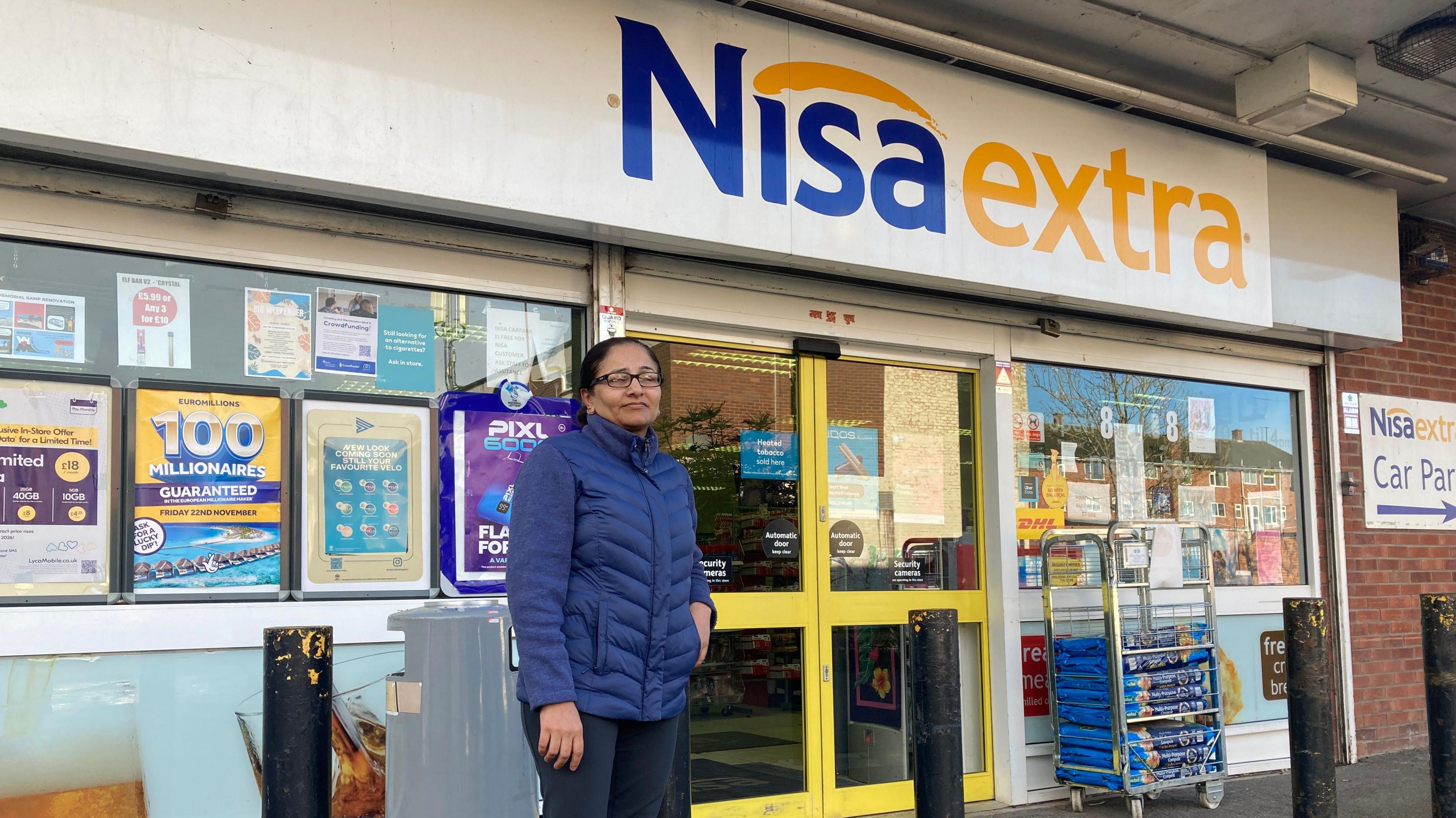 A wide shot of a woman with her hair tied back, wearing glasses and a blue puffer coat standing in front of  a shop with the sign Nisa Extra