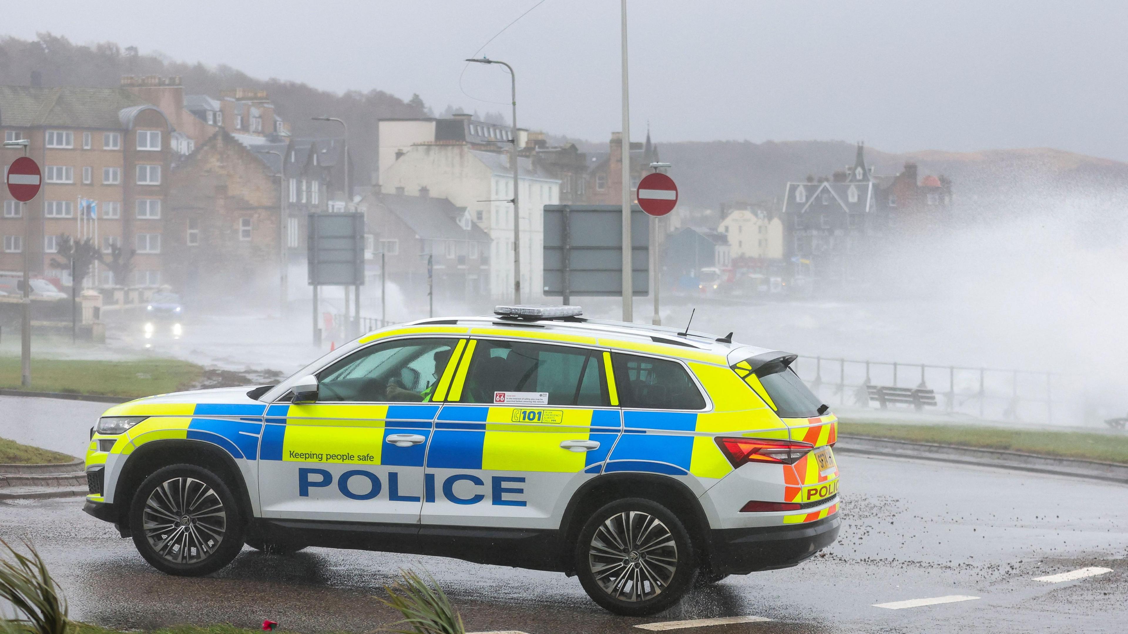 A police car in the foreground with spray from a rough sea and buildings in the background.