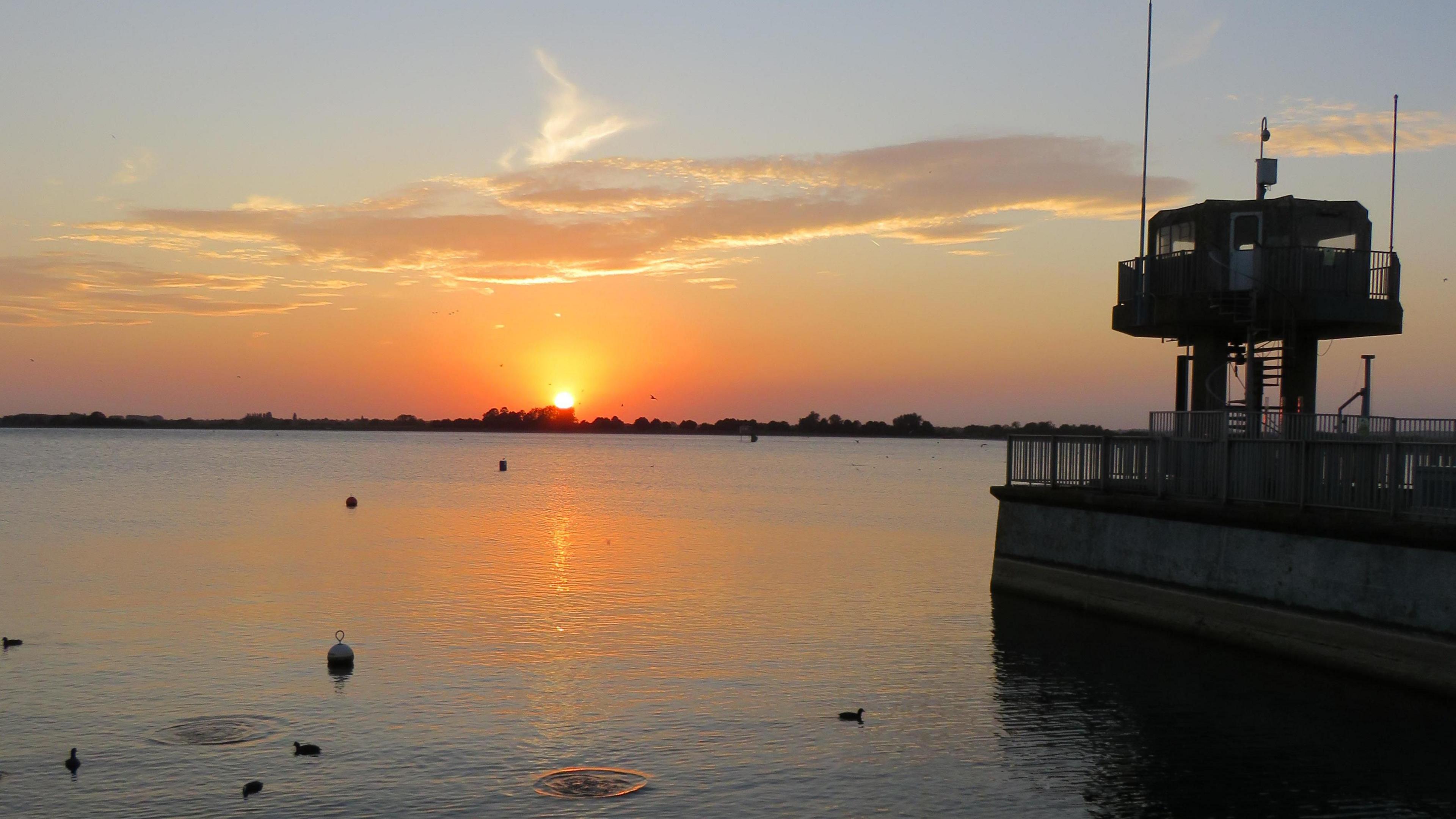 A beautiful sunset over water with ducks on top of the water. A structure can be seen jutting into the water on the right-hand side. The sky is a warm orange with a few white clouds stretching out.