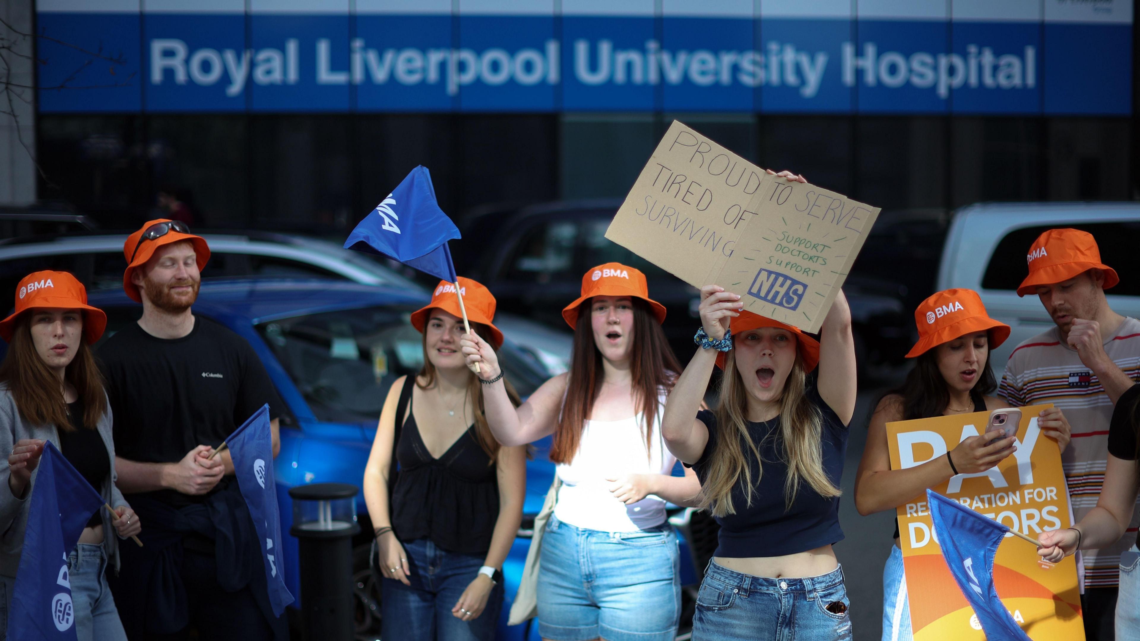 Striking resident doctors hold up placards during a picket outside the Royal Liverpool University Hospital in Liverpool, Britain, 25 July 2025. They wear orange hats and are dressed casually. 
