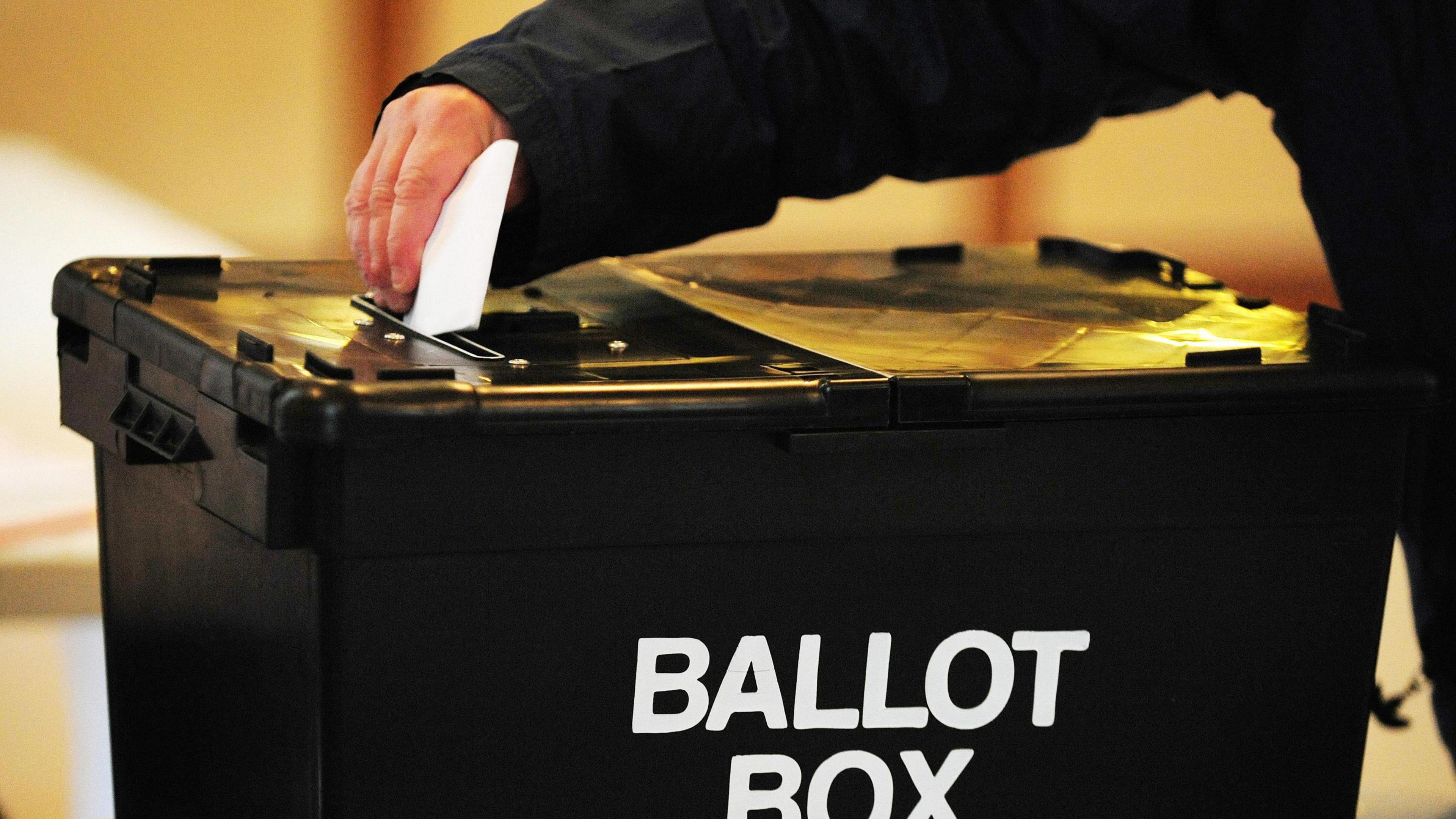 Image shows large black box with BALLOT BOX written on it in white.
There is a hand placing a folded slip of paper into the box. 