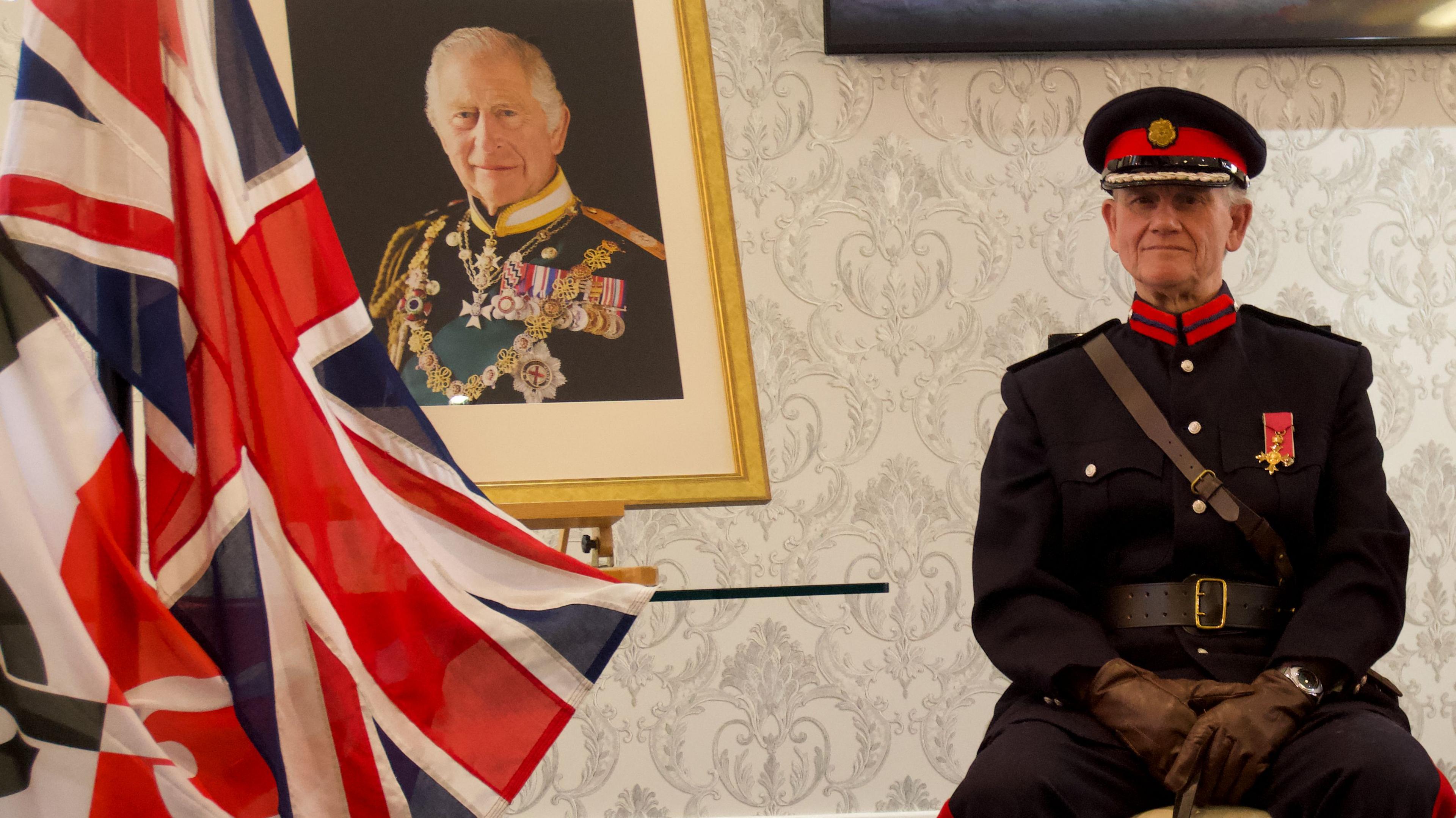 The Black Country flag, the Union flag, a photo of King Charles, wearing military uniform and medals, in a golden frame and a man sitting in navy blue military uniform with brown leather gloves and a navy and red peaked cap 