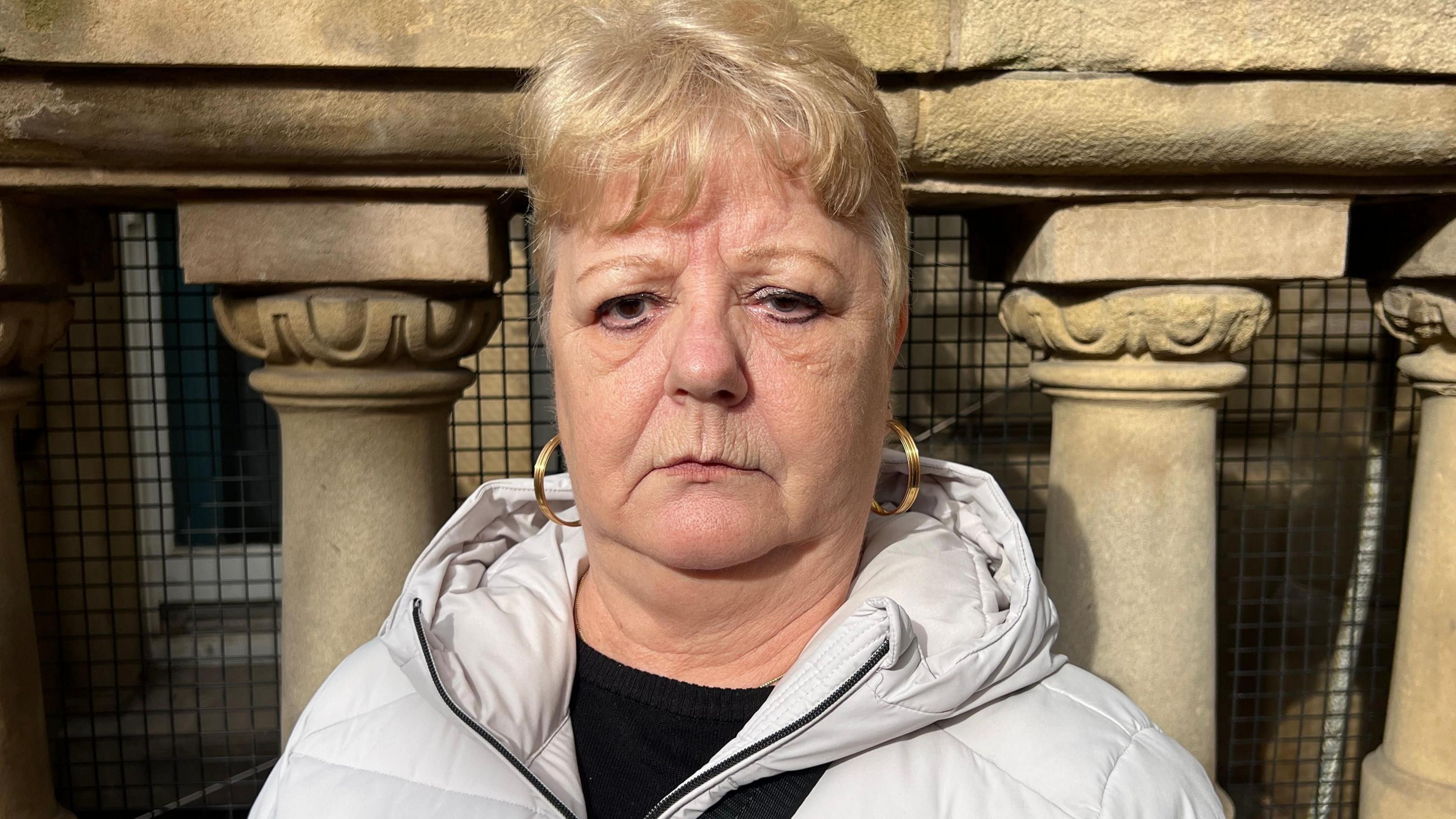 Jayne Harper is standing in front of stone columns out the front of Birmingham City Council House in Victoria Square. She wears a white coat, black top and gold hooped earrings.