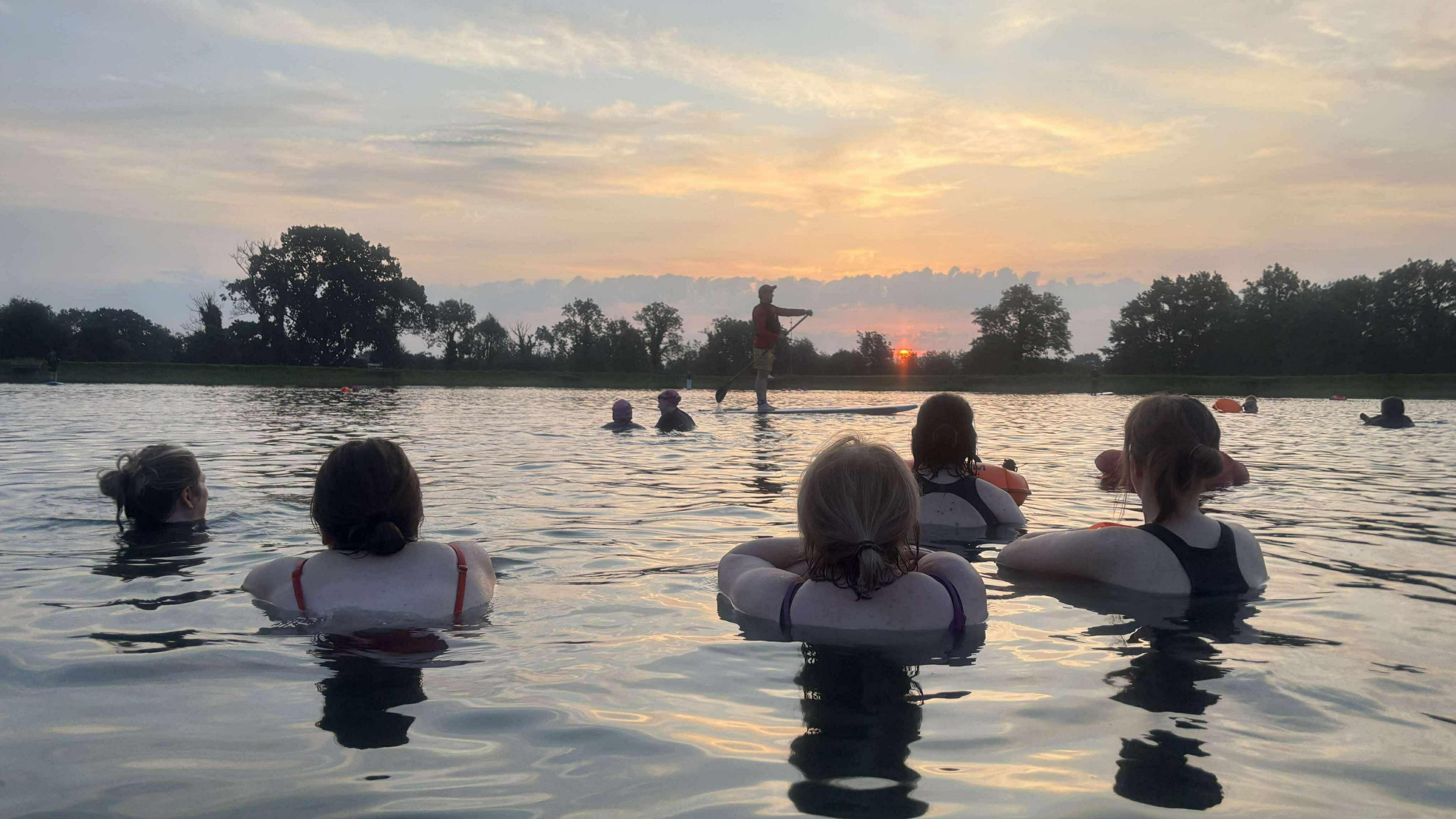 Five women swimming in Moira Lakes early in the morning. The sunrise is visible in the distance. They are all looking away from the camera.