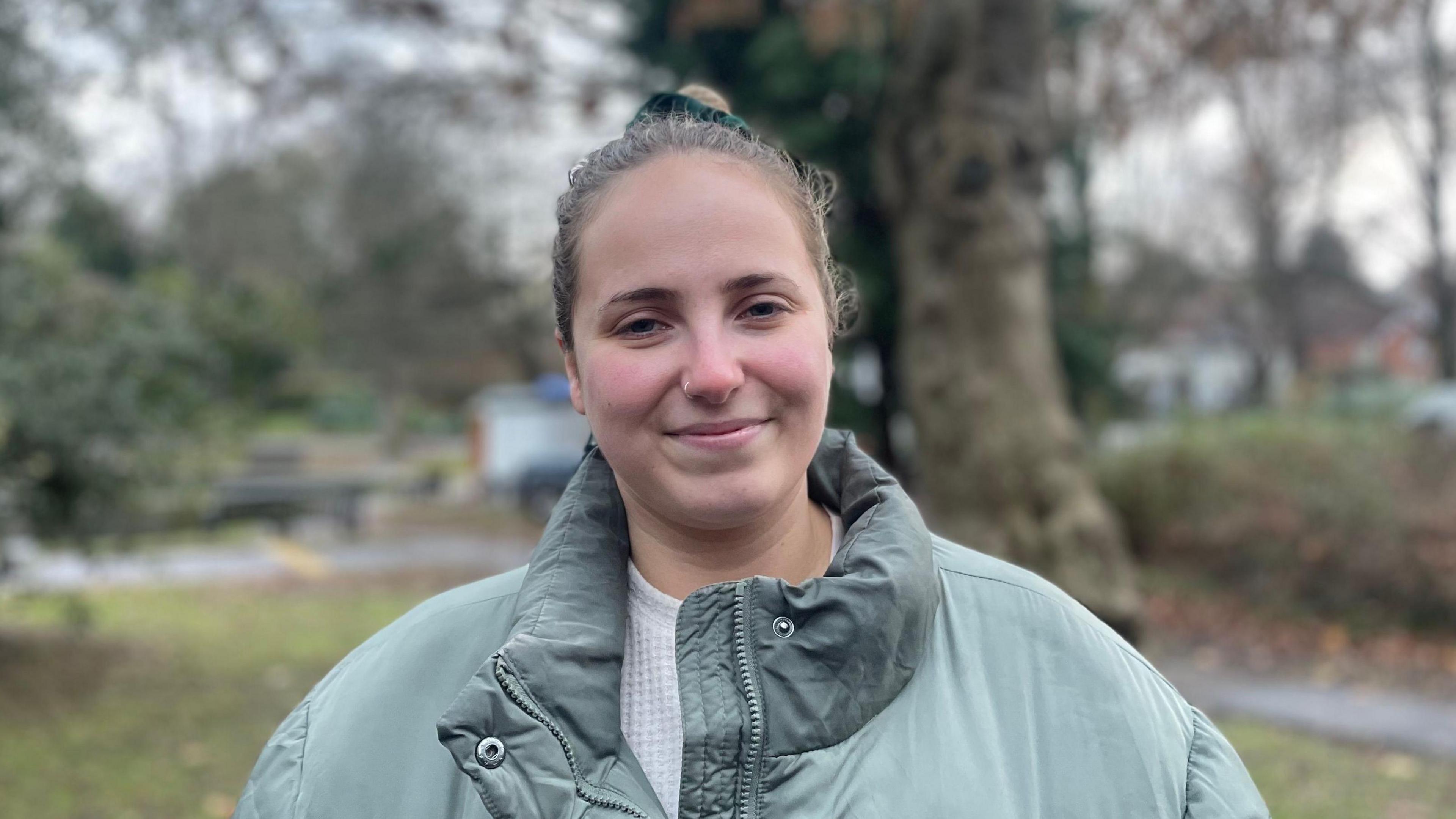 Jessie Crosby is wearing a green coat, smiling and standing in Castle Park. There is a tree behind her. It is a muggy looking day of weather.