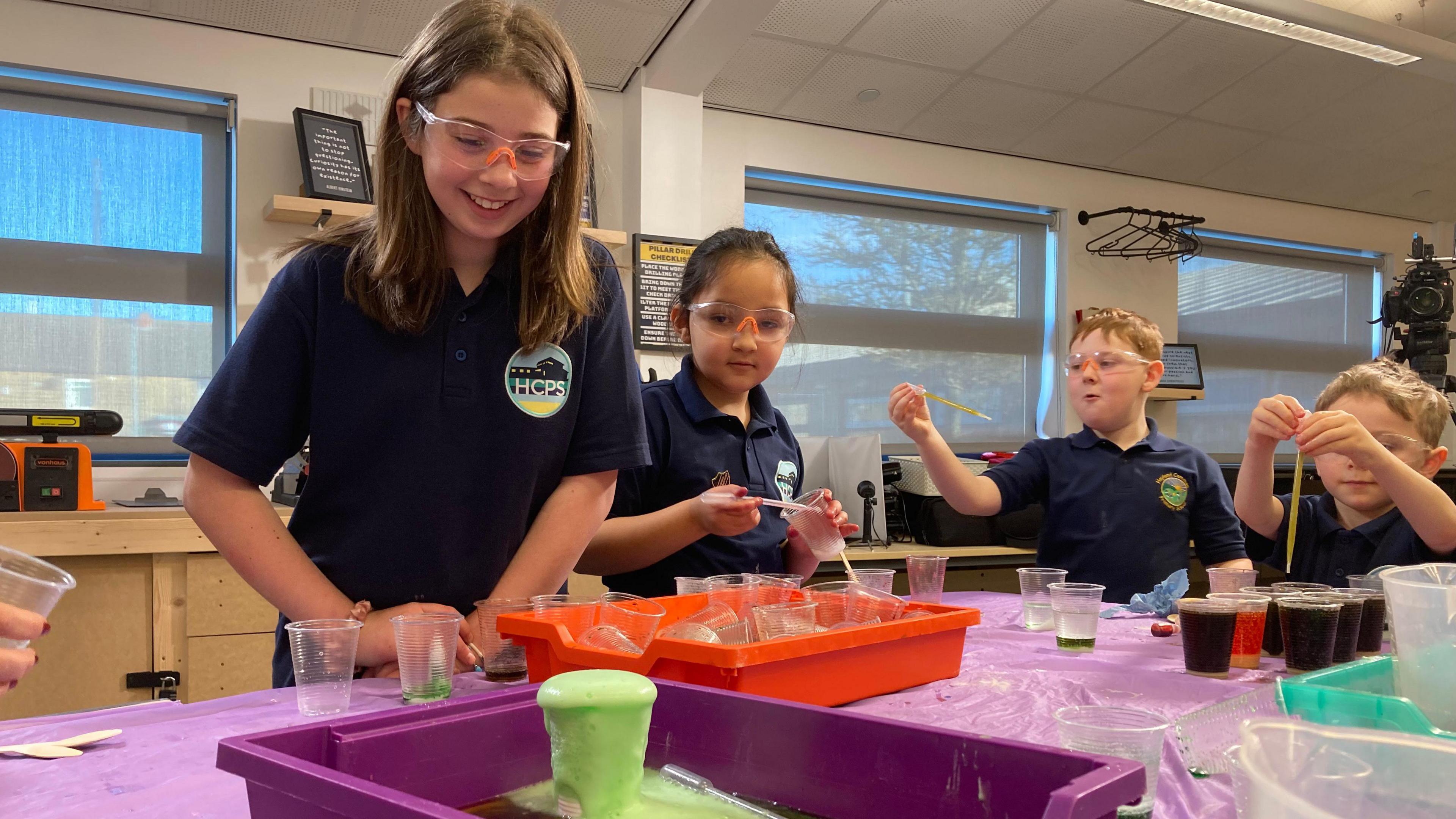 Four children wearing safety goggles can be seen standing around a table. A pile of empty plastic cups have been collected in an orange tray. In a purple tray, a plastic cup stands filled with green, fizzy, foam that pours down the sides. To the right, a little boy stands in front the table using a pipette. He has several plastic cups filled with dark liquid standing in front of him.