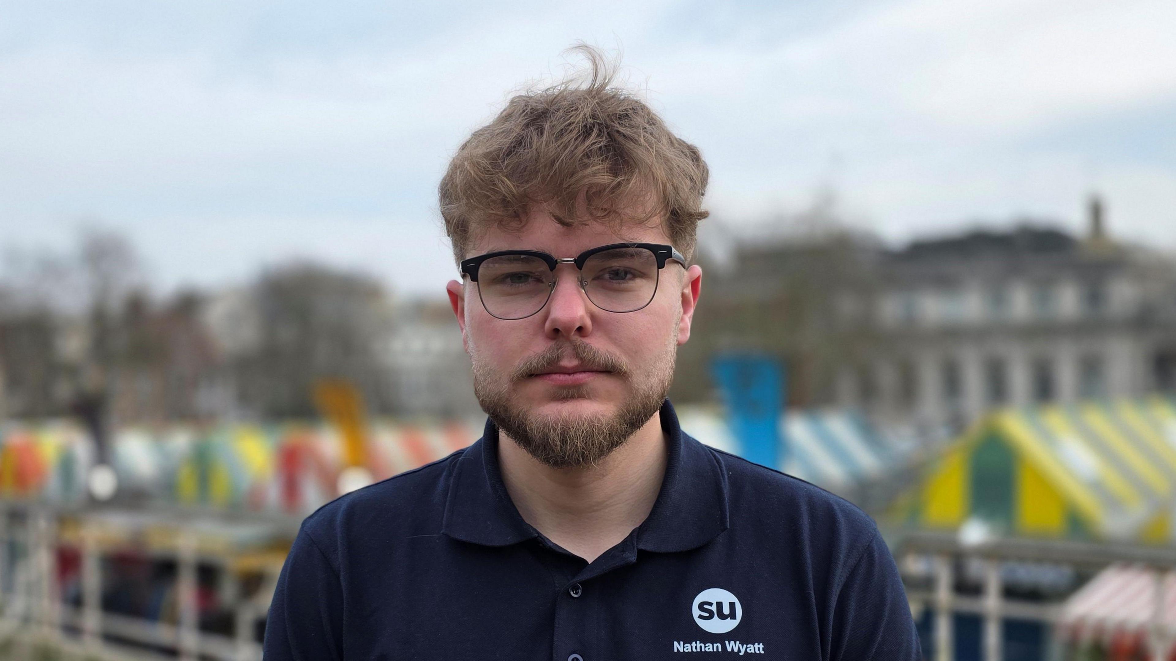 Nathan Wyatt is wearing a dark blue t-shirt with the student union logo and his name on it. He's wearing glasses. Behind him, we can see Norwich marketplace.