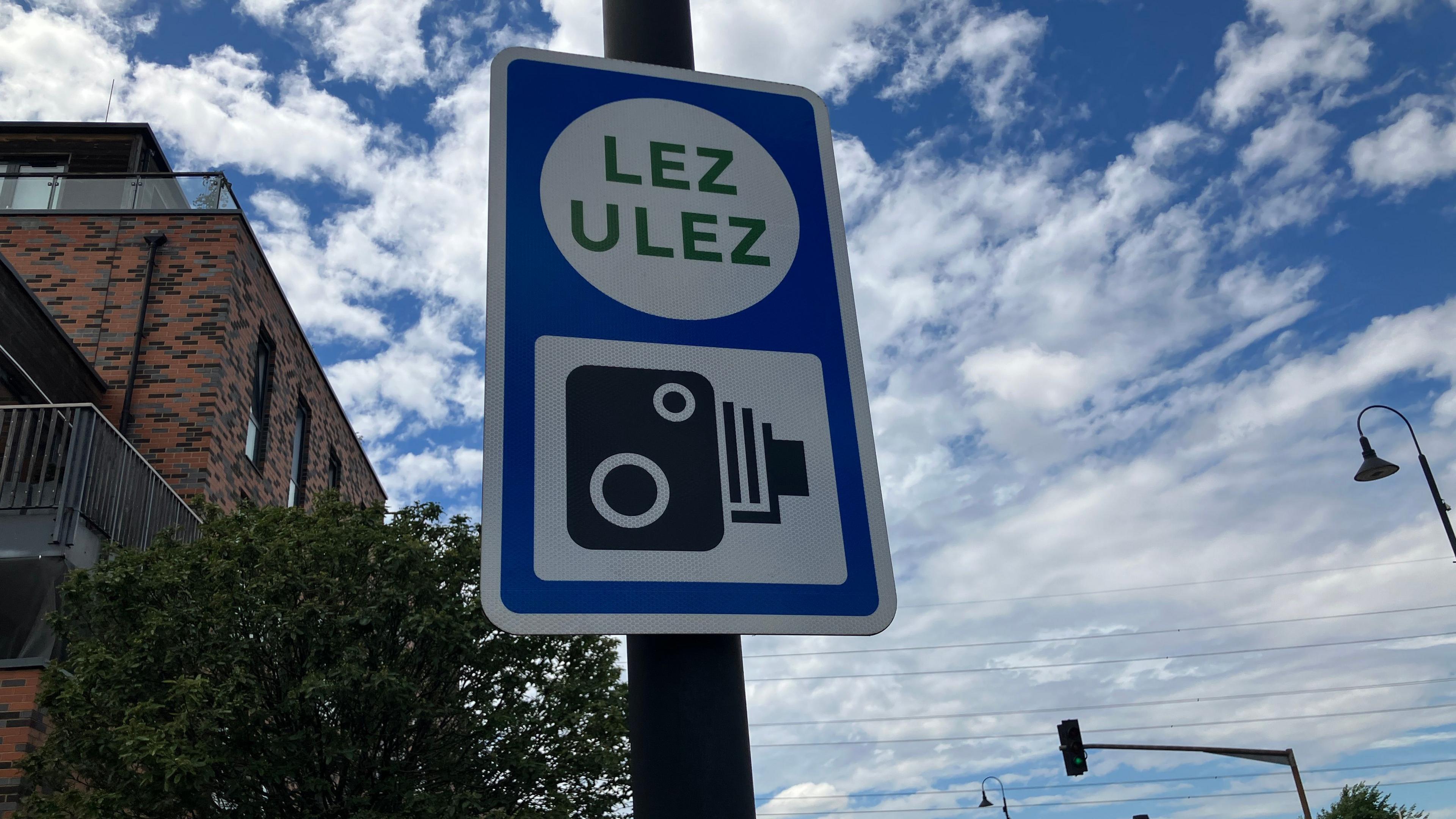 A blue roadside sign displaying the LEZ and ULEZ symbols is mounted on a pole beside a road. A brick building, trees, streetlights, traffic lights and moving vehicles are visible beneath a partly cloudy sky.