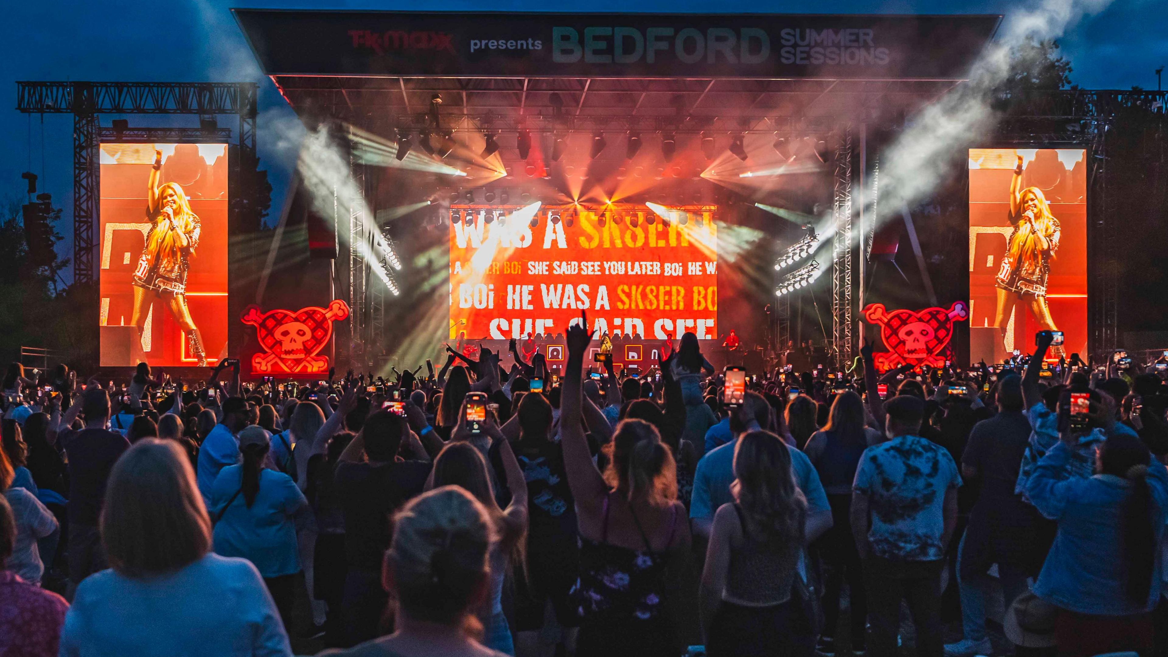 Avril Lavigne performing in Bedford Park in 2024, showing a stage, with screens, her in the middle of the stage, audience members in front, and stage lights. 