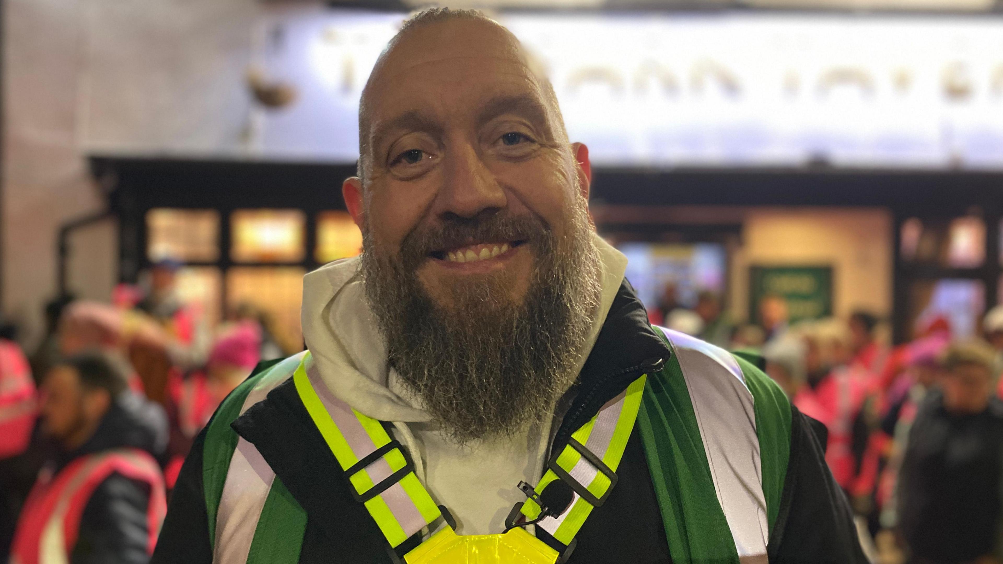 Carl Ackerman is wearing a green high-vis jacket and a grey hoodie. He is standing in front of crowds of people wearing pink high-vis jackets.