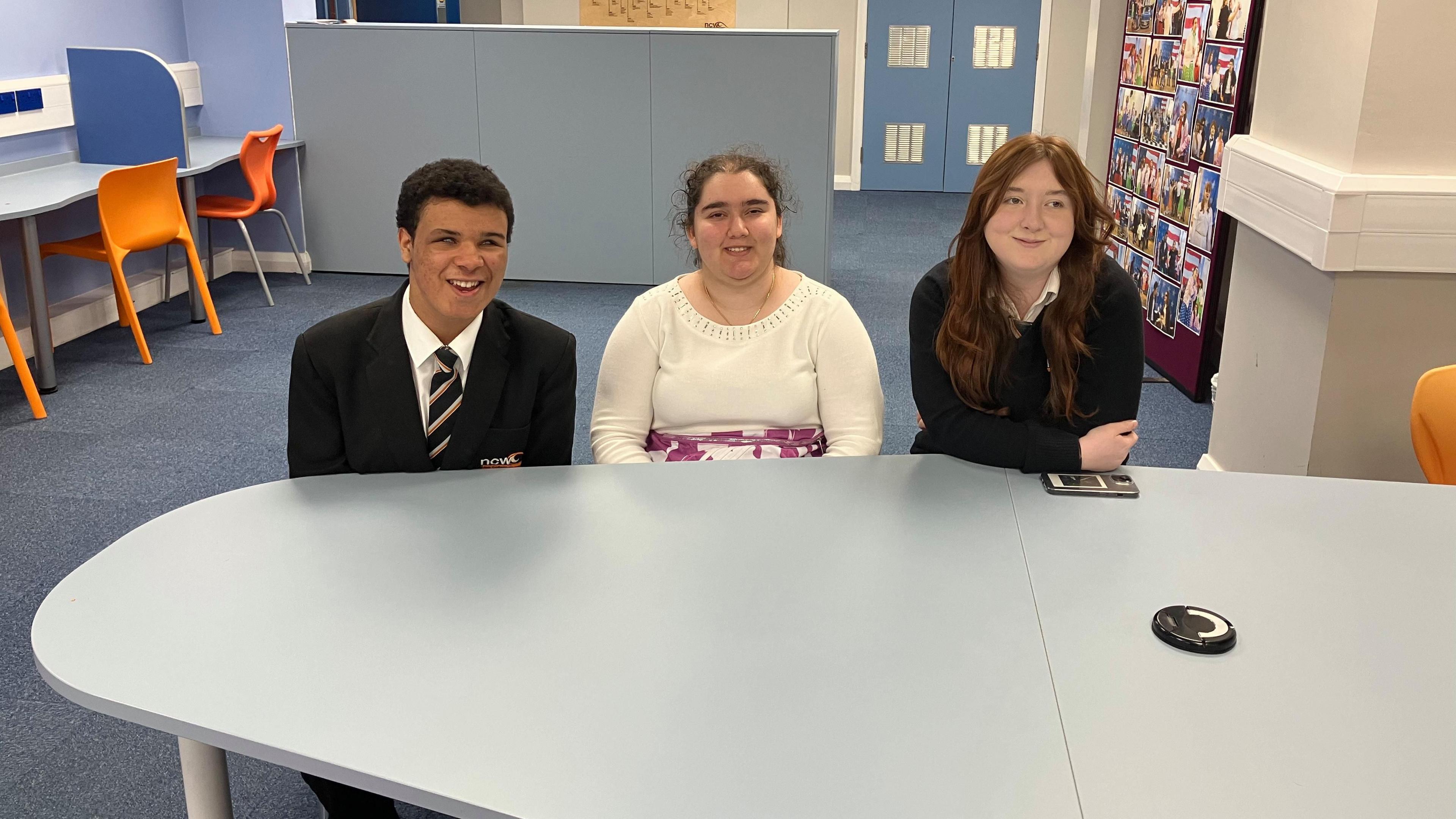 Three teenagers sit at a large desk in a classroom. On the left, a boy with short dark hair is wearing a dark suit with a white shirt and a striped tie, a girl in the middle with tied-back dark curly hair is wearing a cream jumper and on the right, a girl with long dark red hair is wearing a white blouse under a black jumper. 