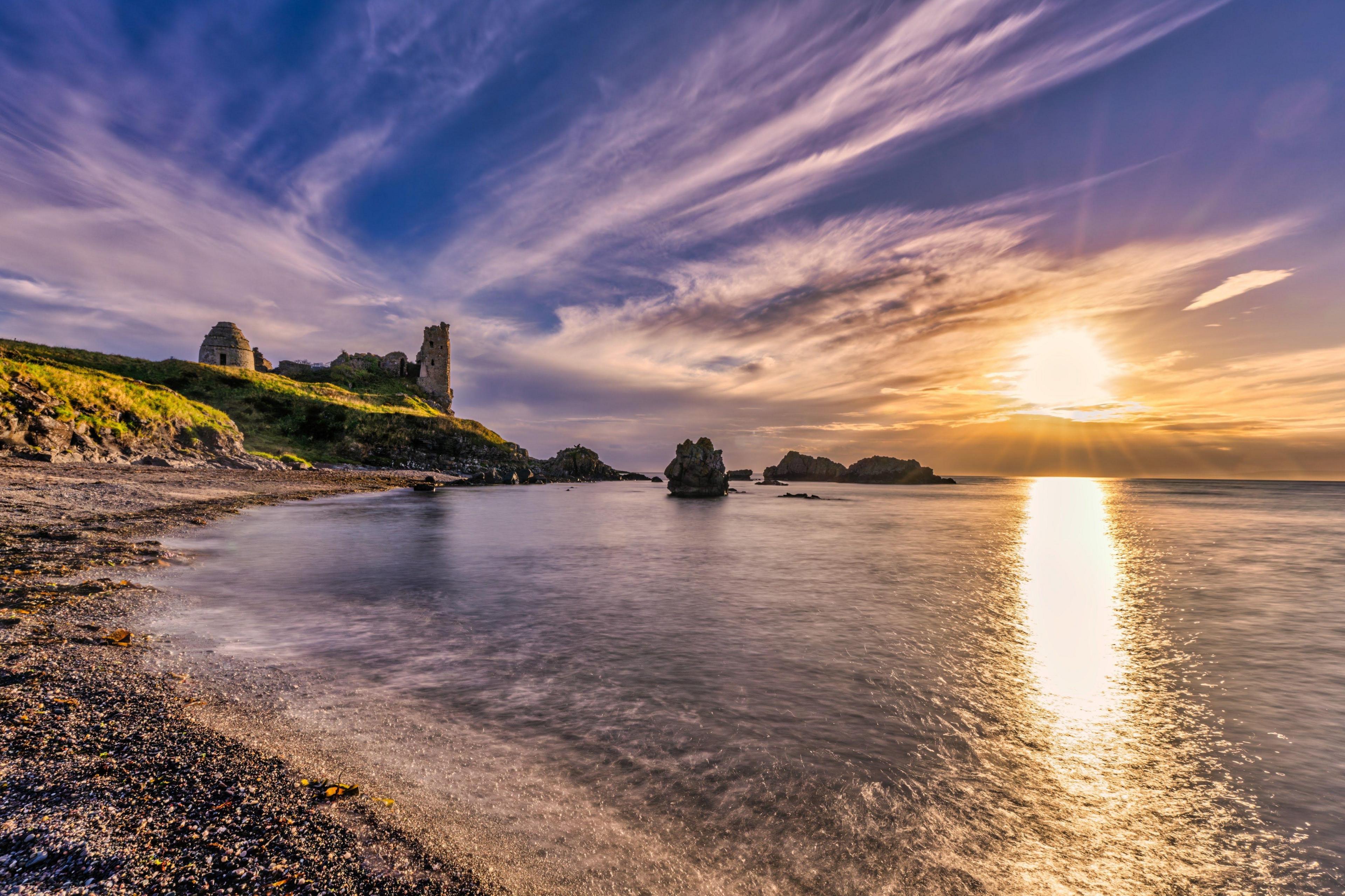 On the left are castle ruins on a grassy hillside which meets the waters edge. The sun is going down and reflecting on the sea. There are many wispy clouds in the sky.