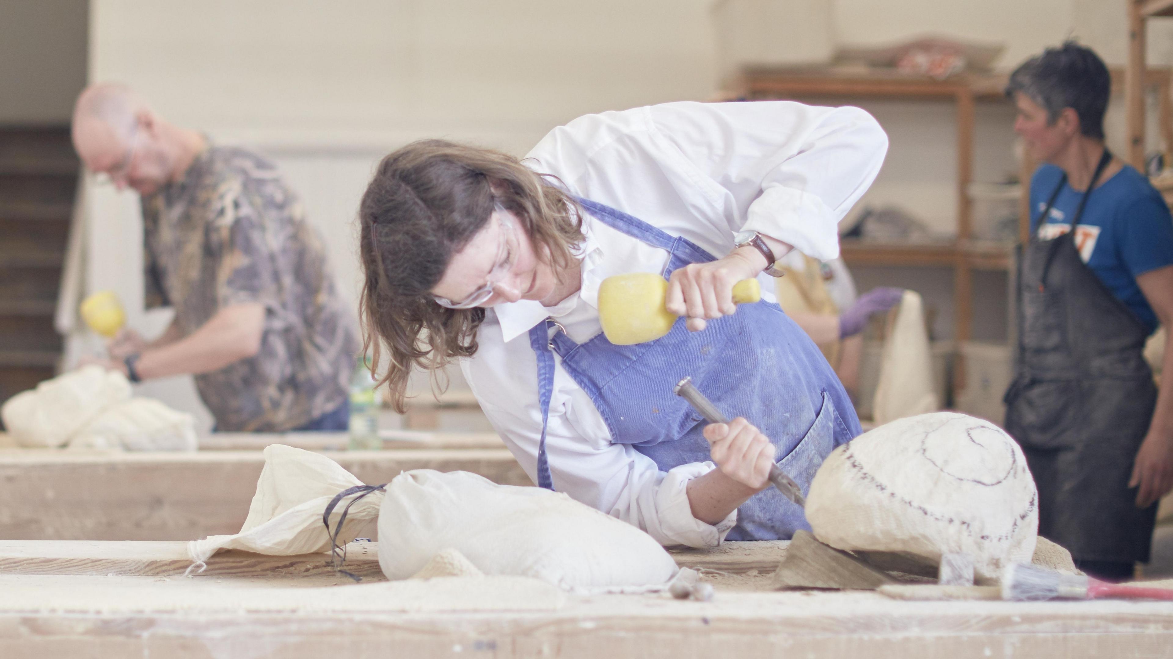 A woman carving stone in a workshop. She is wearing a blue apron over white overalls and protective glasses. In the background there are two other people, one is also carving a stone and the other is standing to the side. 