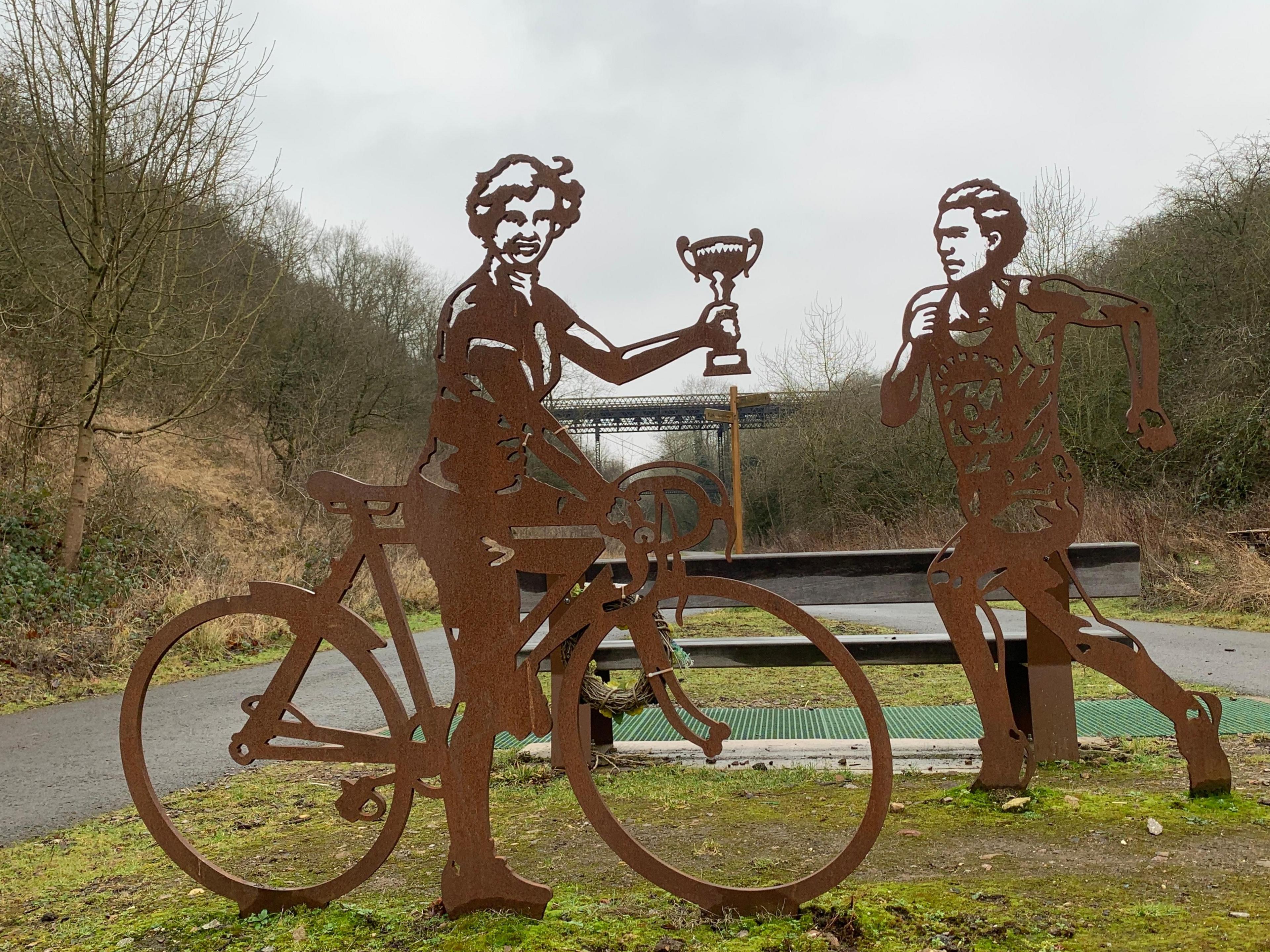 Two weathered cut-out steel sculptures, featuring a woman on a bike holding a trophy aloft and a man running. They are between two lanes of a tarmac cycle track, with old railway embankments on either side and a bridge visible in the background.  