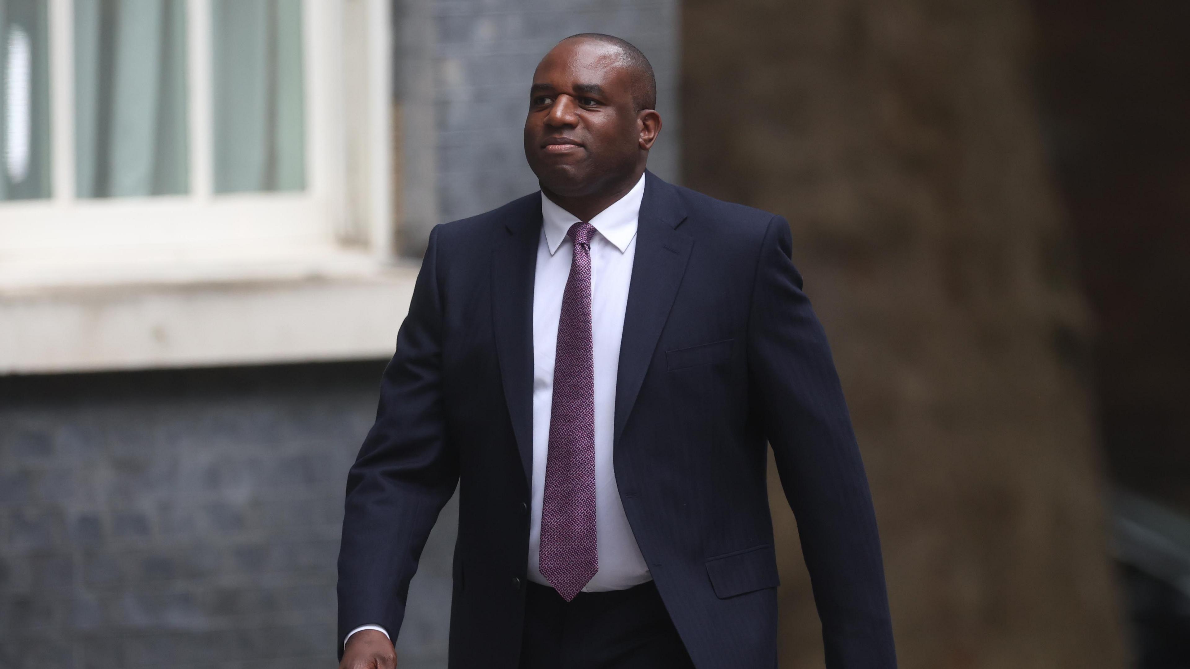 David Lammy walks down Downing Street, wearing a navy blue suit and pale purple tie ign Secretary