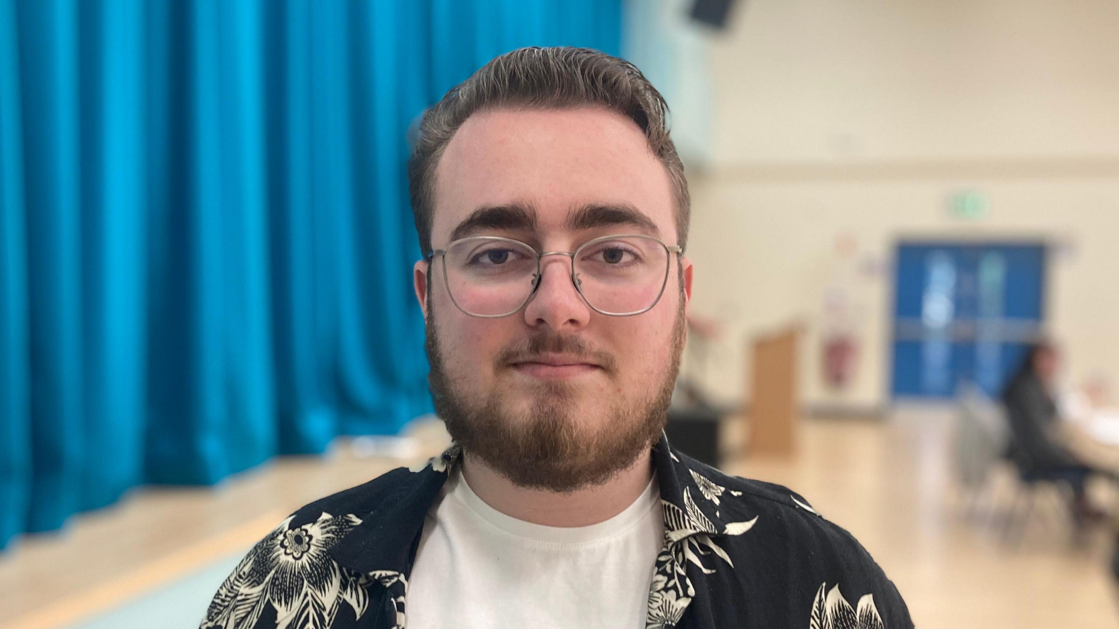 Max has short brown hair and brown facial hair he is wearing silver glasses, a white t-shirt and a black shirt with white flowers on it.