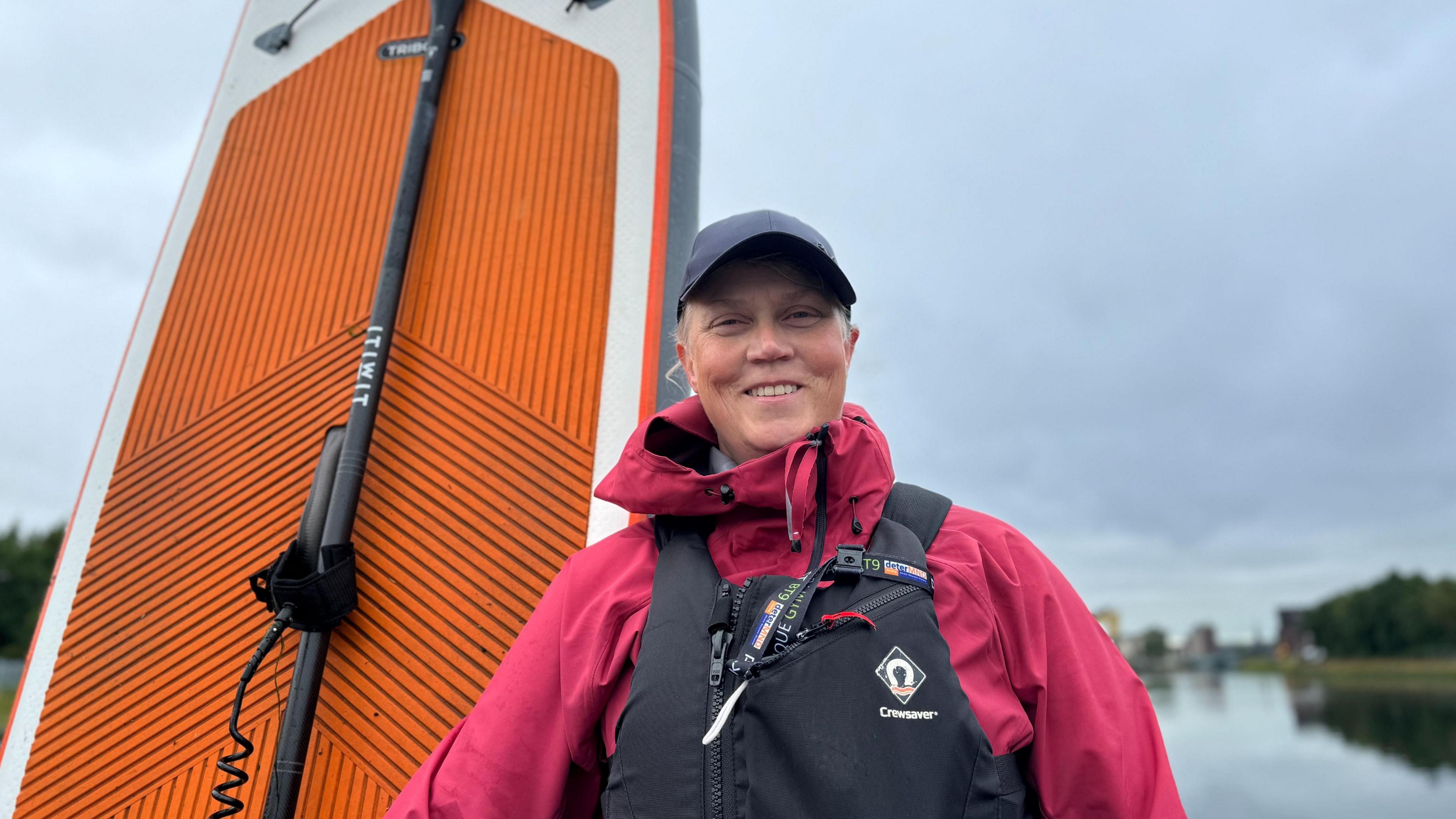 Woman standing in front of a body of water wearing pink waterproof coat and black life jacket holding a stand-up paddleboard