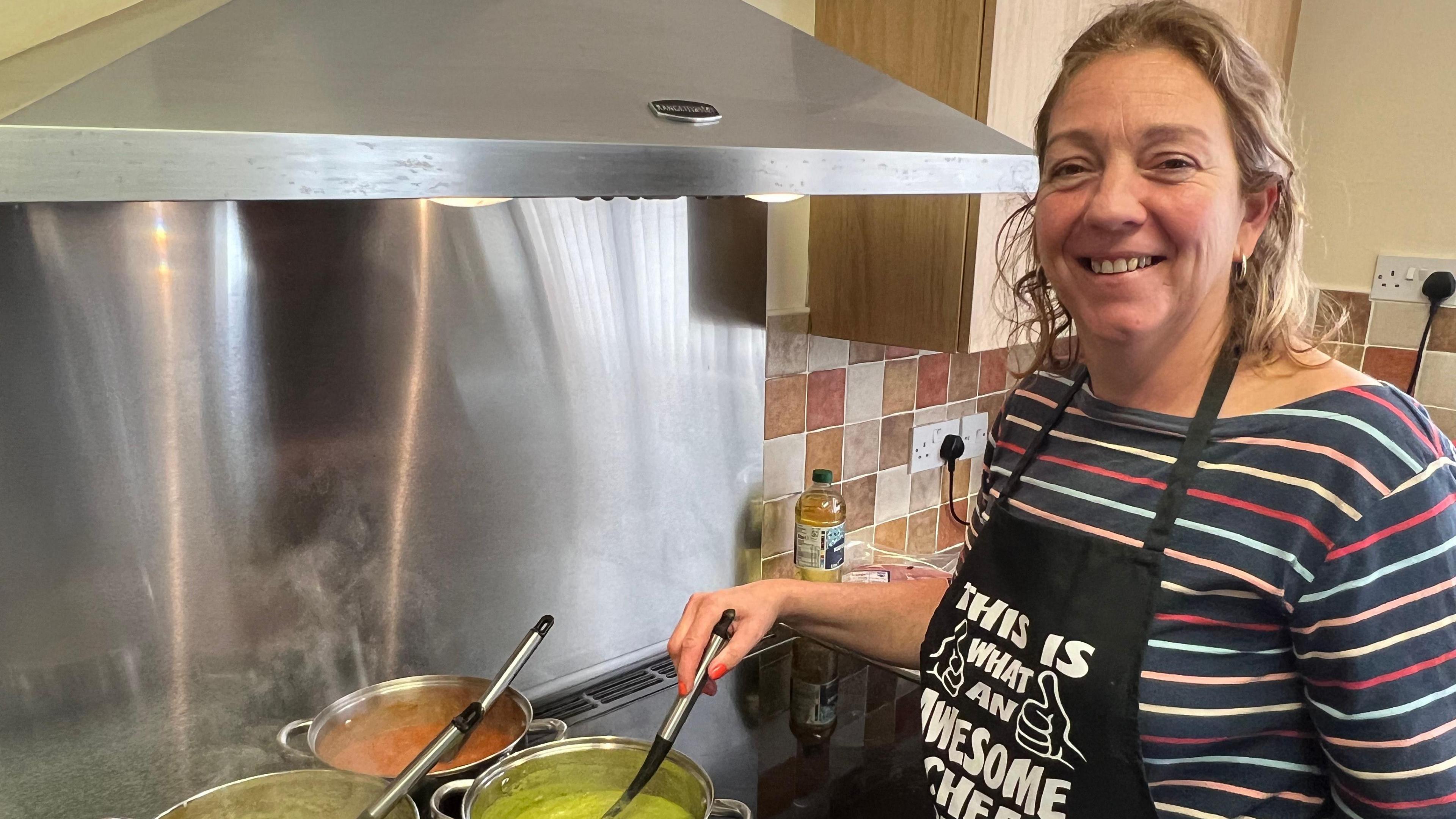 Sam Warren. She has brown short curly hair and is wearing a striped top under a black apron. She is standing at a hob which has three sauce pans on it. She is mixing the contents of one pot, while looking at the camera and smiling. 