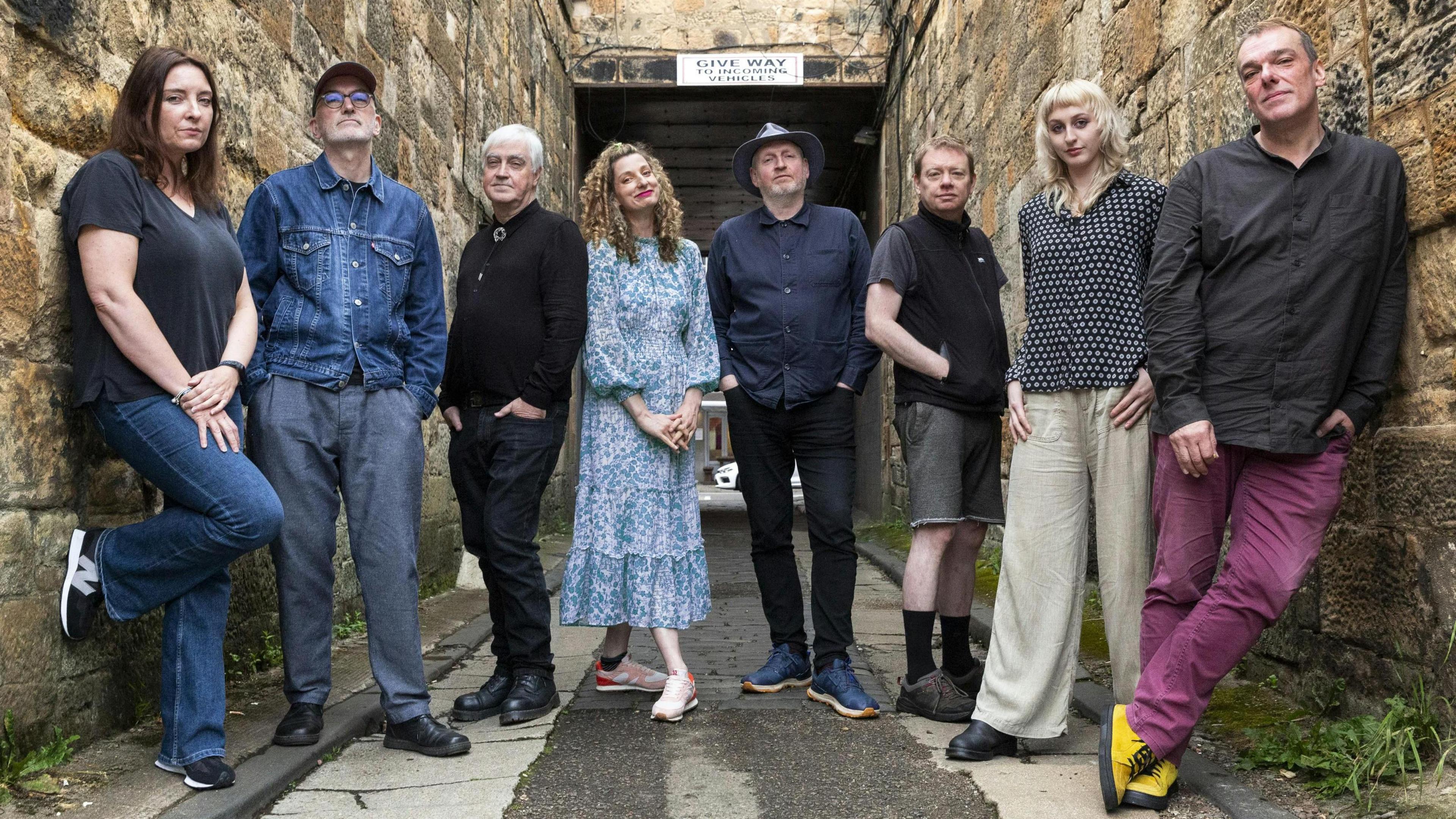 The eight members of the Tenementals, standing in an alleyway while looking straight into the camera. They are all dressed casually, with some members leaning against the walls of the alley.