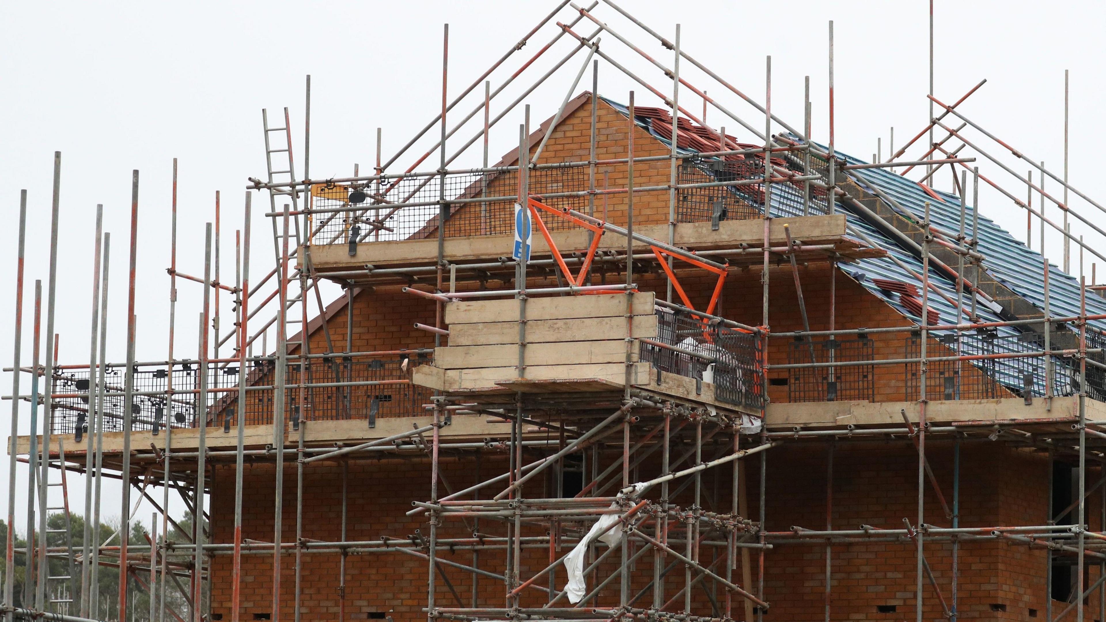 Scaffolding surrounds a two-storey red-brick house under construction. Tiles are stacked on its roof.