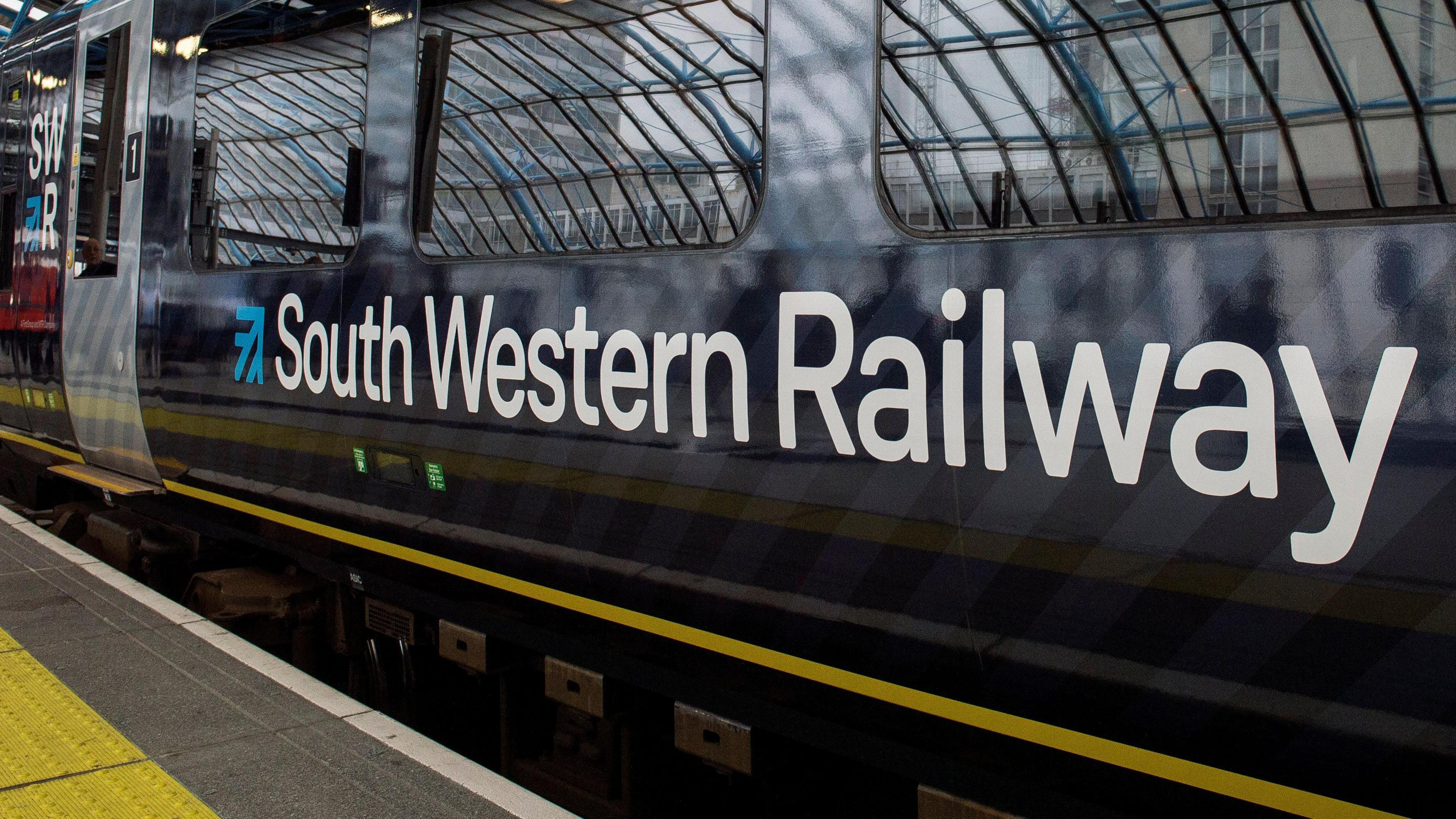 A file image showing the white and blue South Western Railway logo on a train with a yellow and light and dark blue livery as it is pulled into the platform