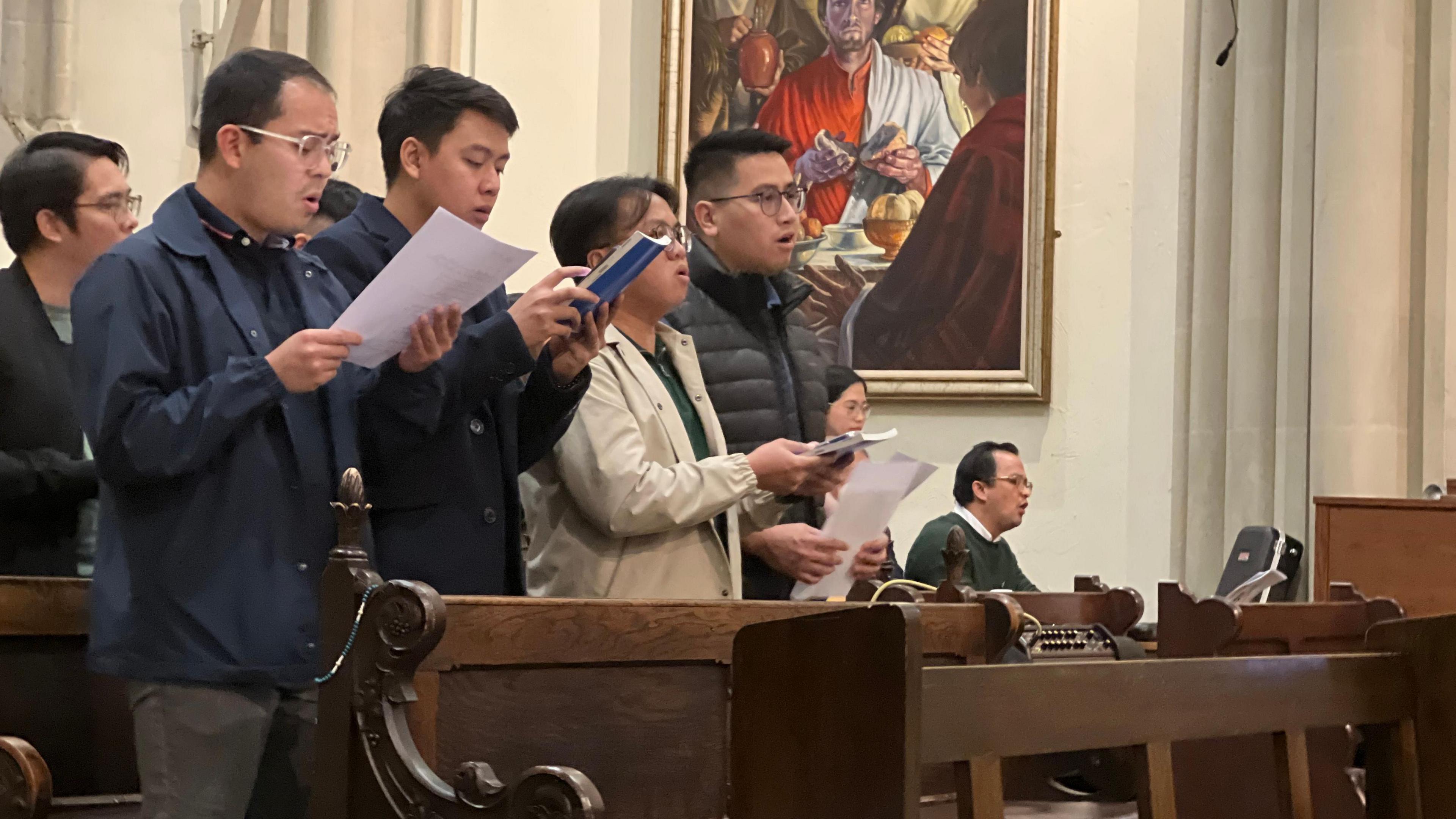 A group of men sign with sheets in front of them. They are standing behind wooden pews in the church with a church painting behind them. They are wearing coats and behind them more people are singing.