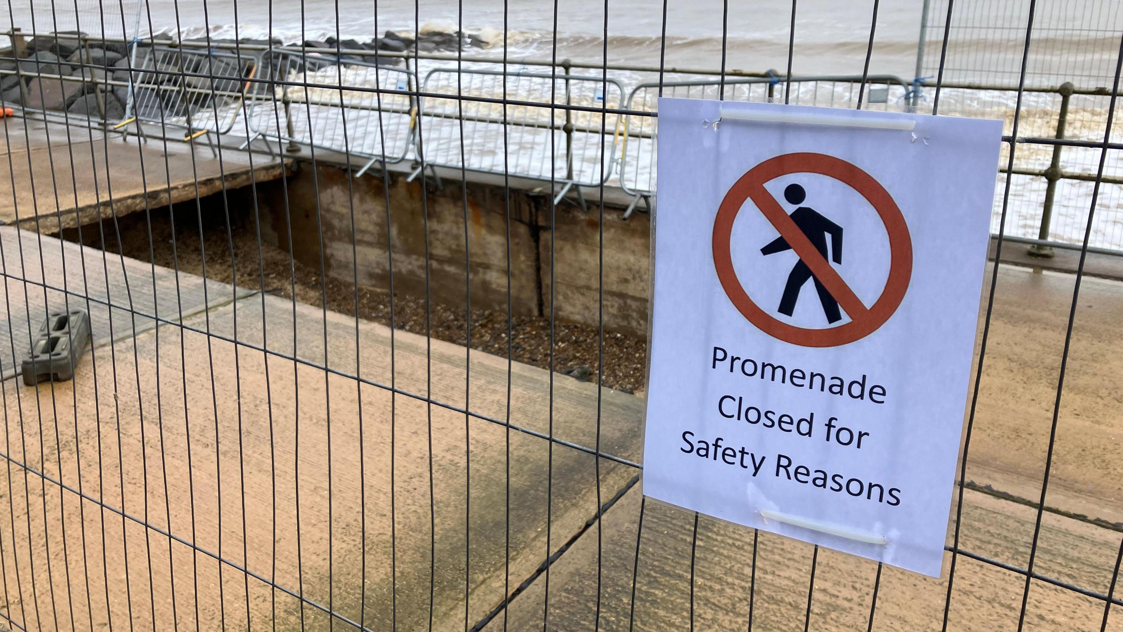 A large rectangular-shaped hole surrounded by metal fencing in the seafront walkway and a sign reading Promenade Closed for Safety Reasons