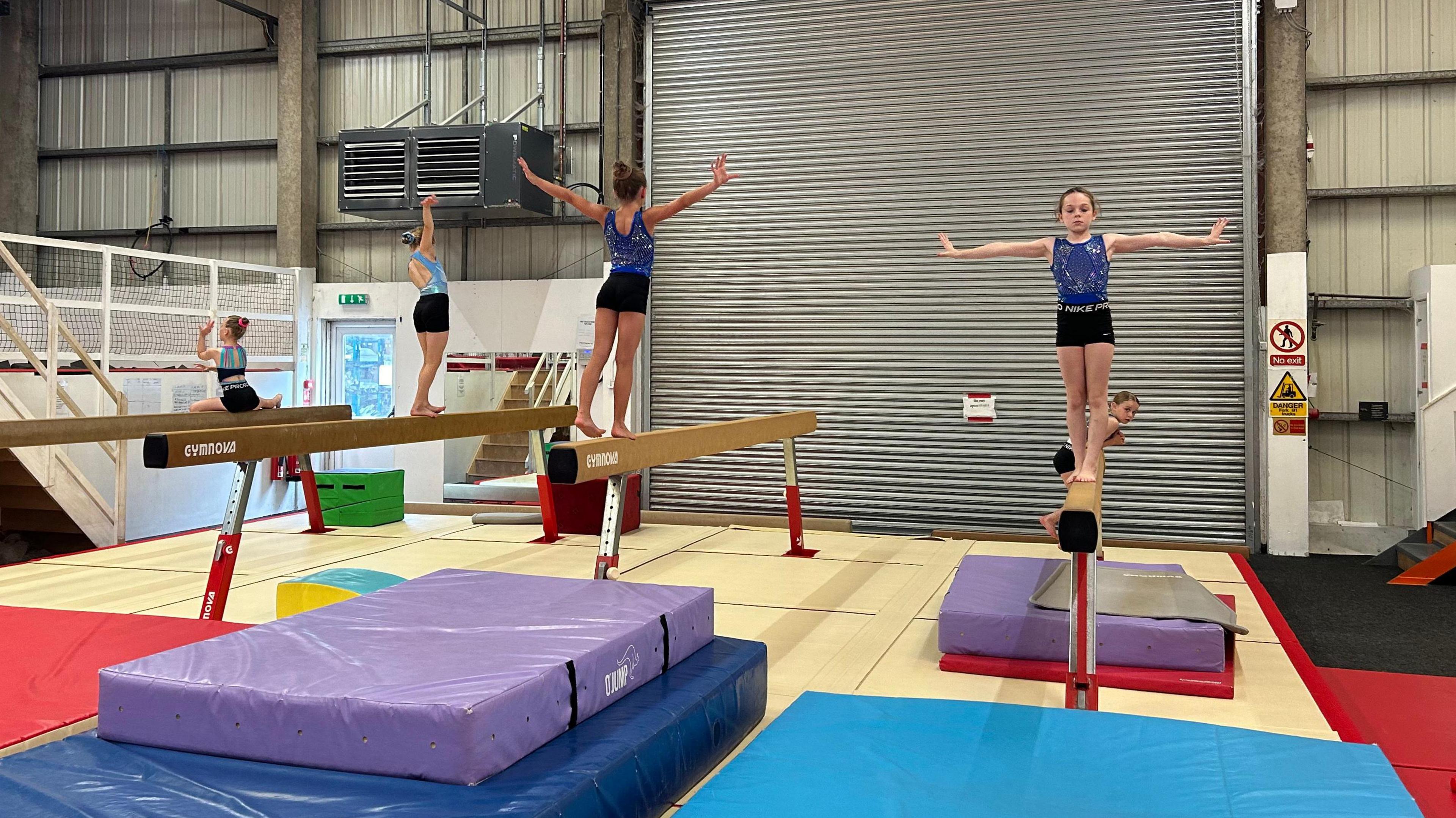 A group of girl gymnasts performing moves on the balance beam with crash mats placed at the end of the equipment