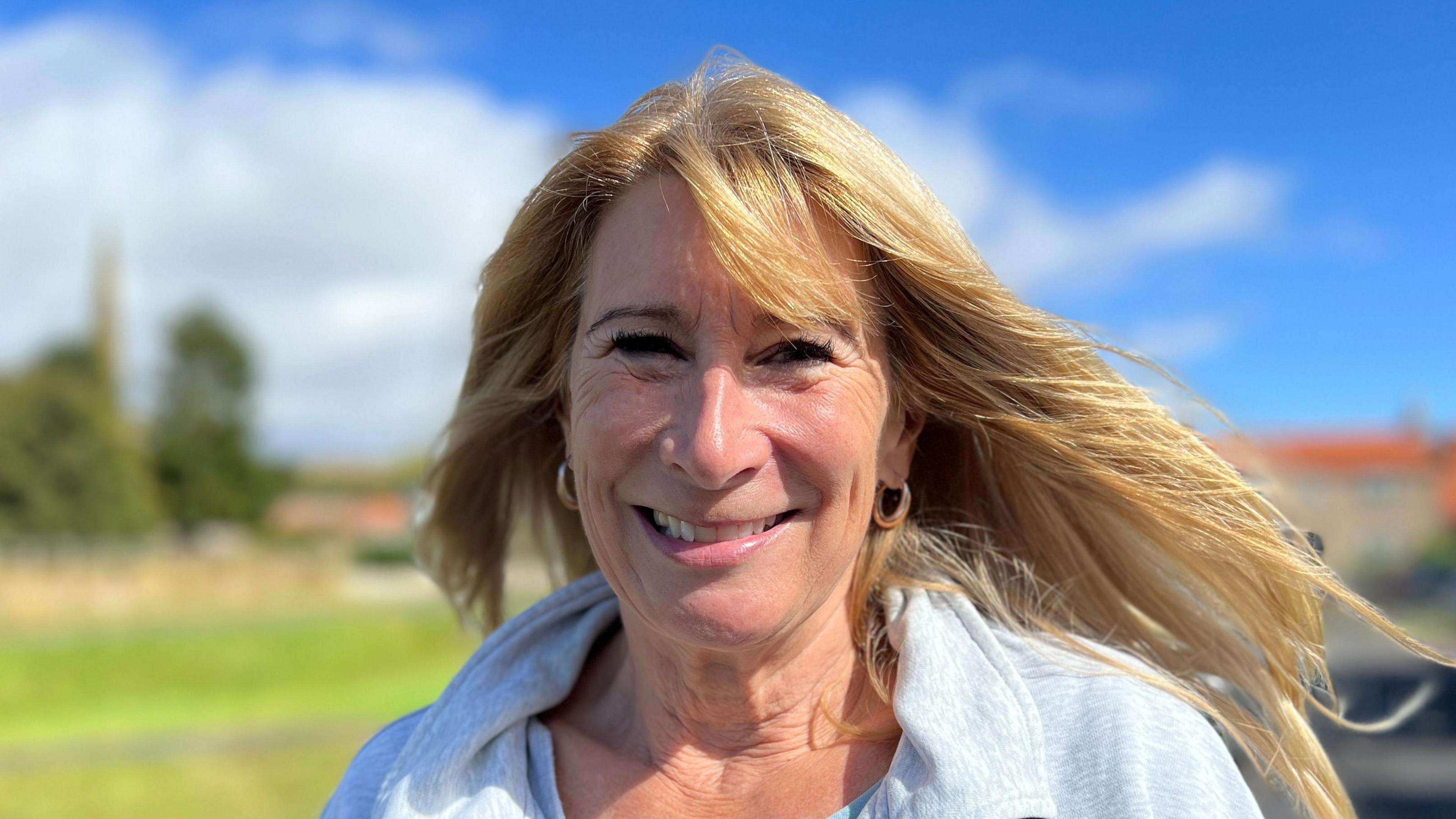 Ingrid Haywood has long blonde hair which has been blown by the wind. A smile has filled her face and her cheeks are rosy. She has a grey jumper on. She's standing in front of several buildings and the village church, the background behind her has been blurred.