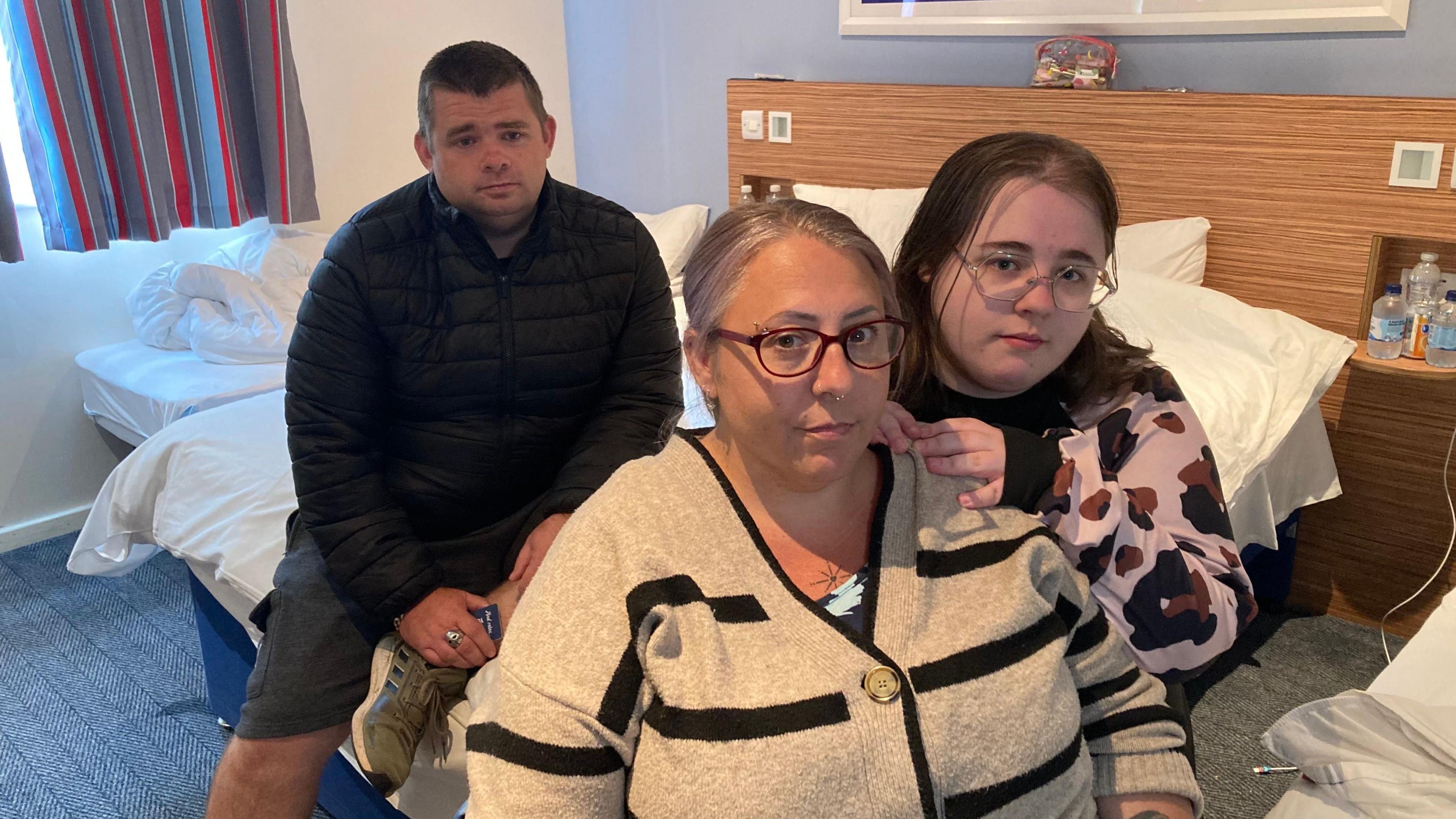 A man and two women sat on a bed in a hotel room. Both women are wearing glasses and all three are looking towards the camera. There is a window and blue and red curtains in the background.
